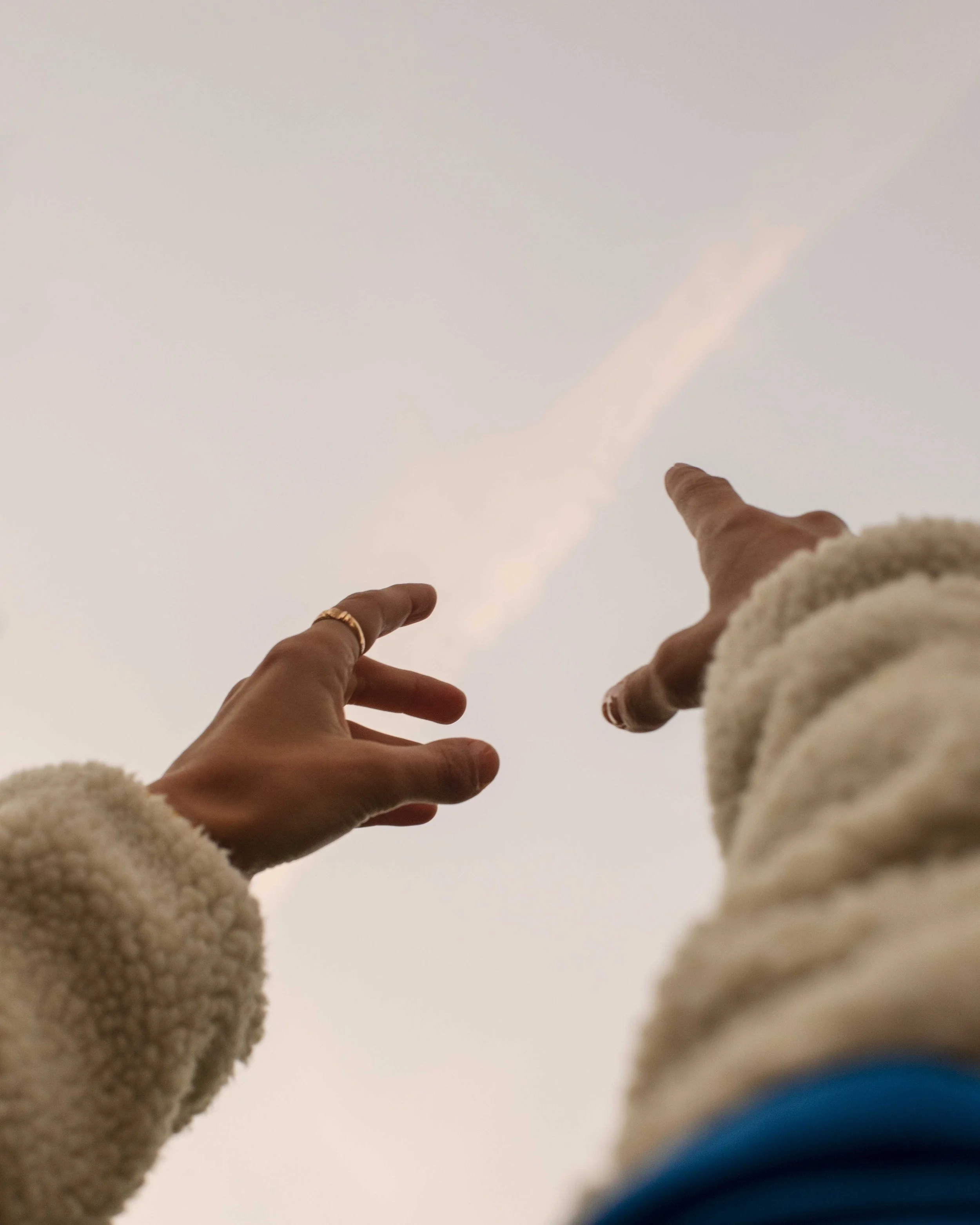 Two hands reaching up towards the sky, one with a ring, in a cozy cream-colored sweater, with a cloudy sky background.