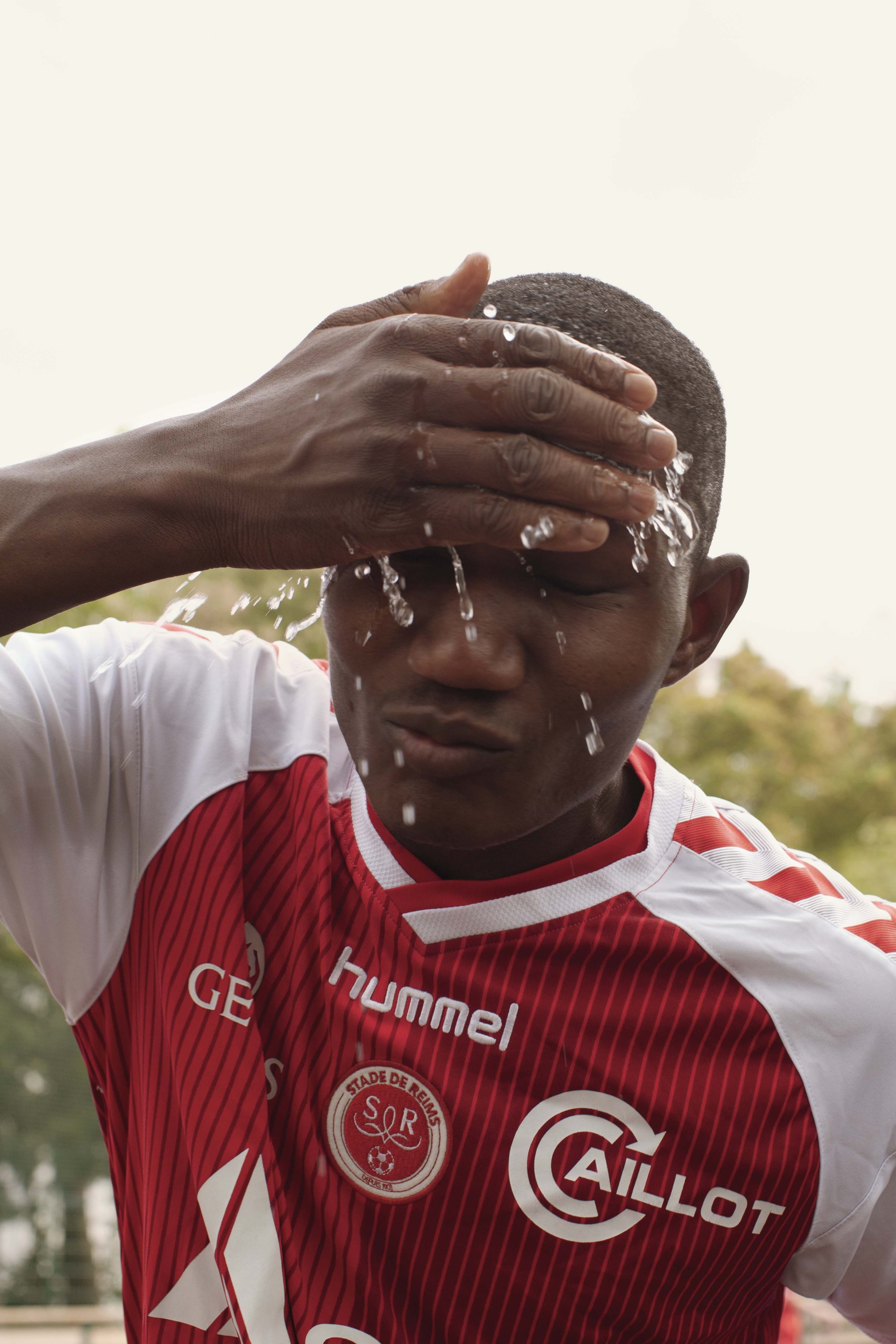 A male soccer player in a red and white jersey rinses his face with water during a game, with trees in the background.