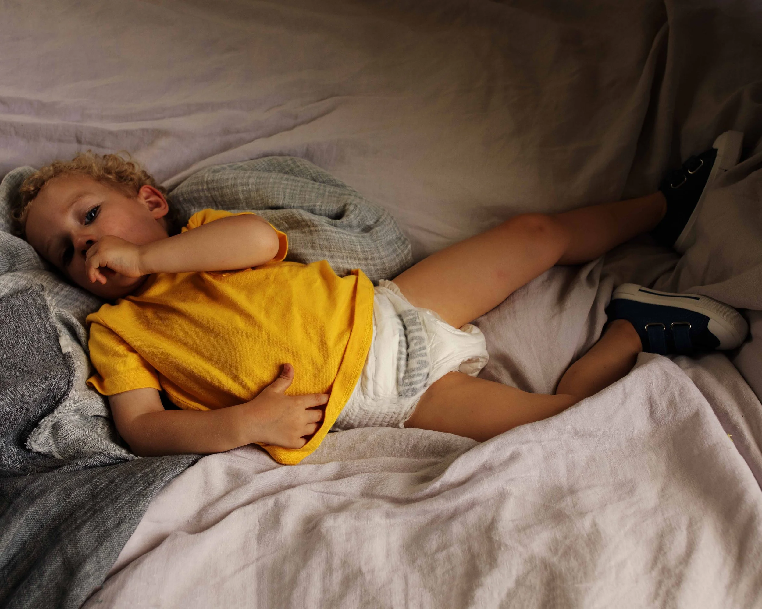 A young boy with curly blond hair lying on a bed, wearing a yellow shirt, white diaper, and sneakers, resting with one hand near his face and the other on his stomach.