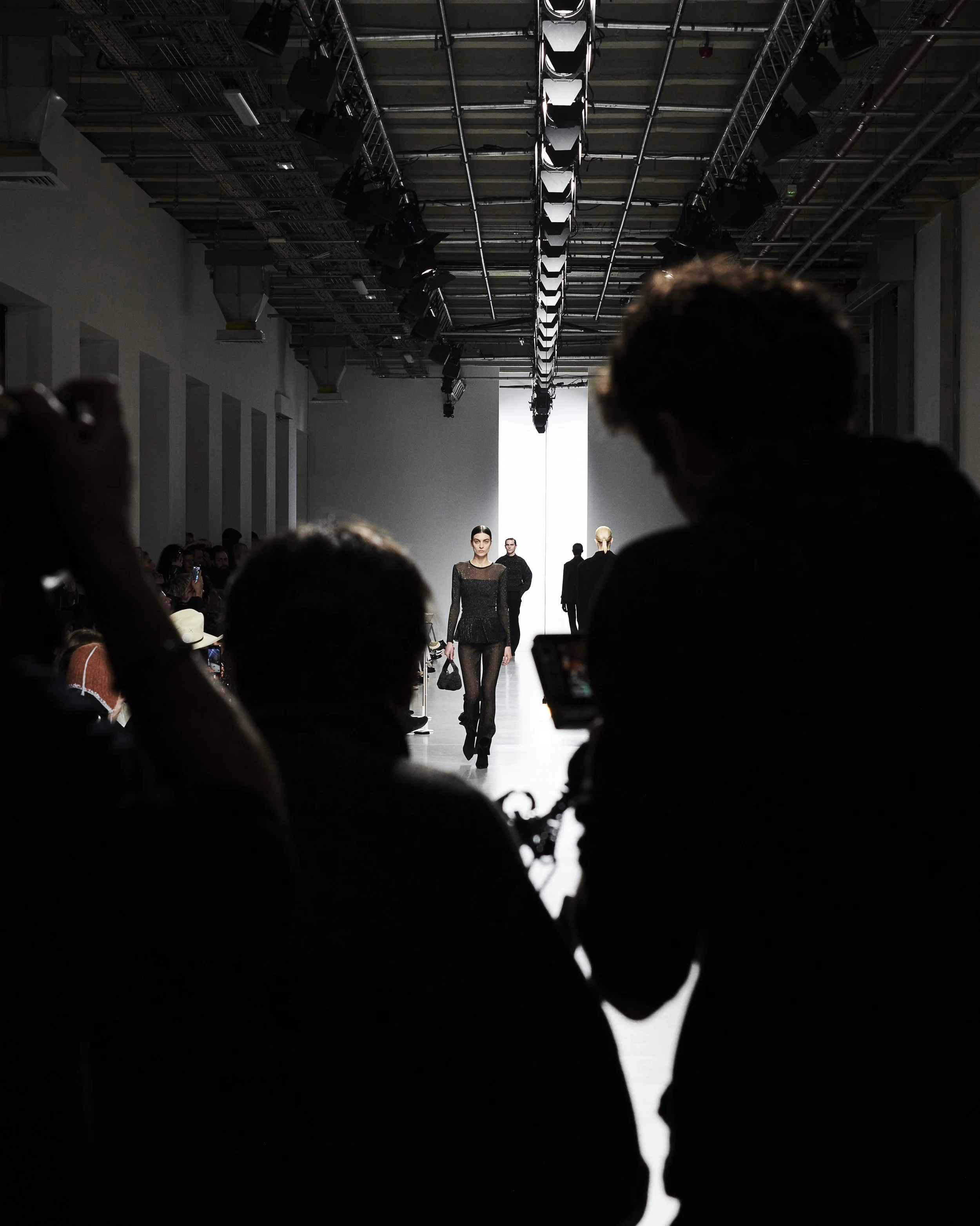 Models walk down the runway during a fashion show, with an audience taking photos and watching in silhouette in the foreground.