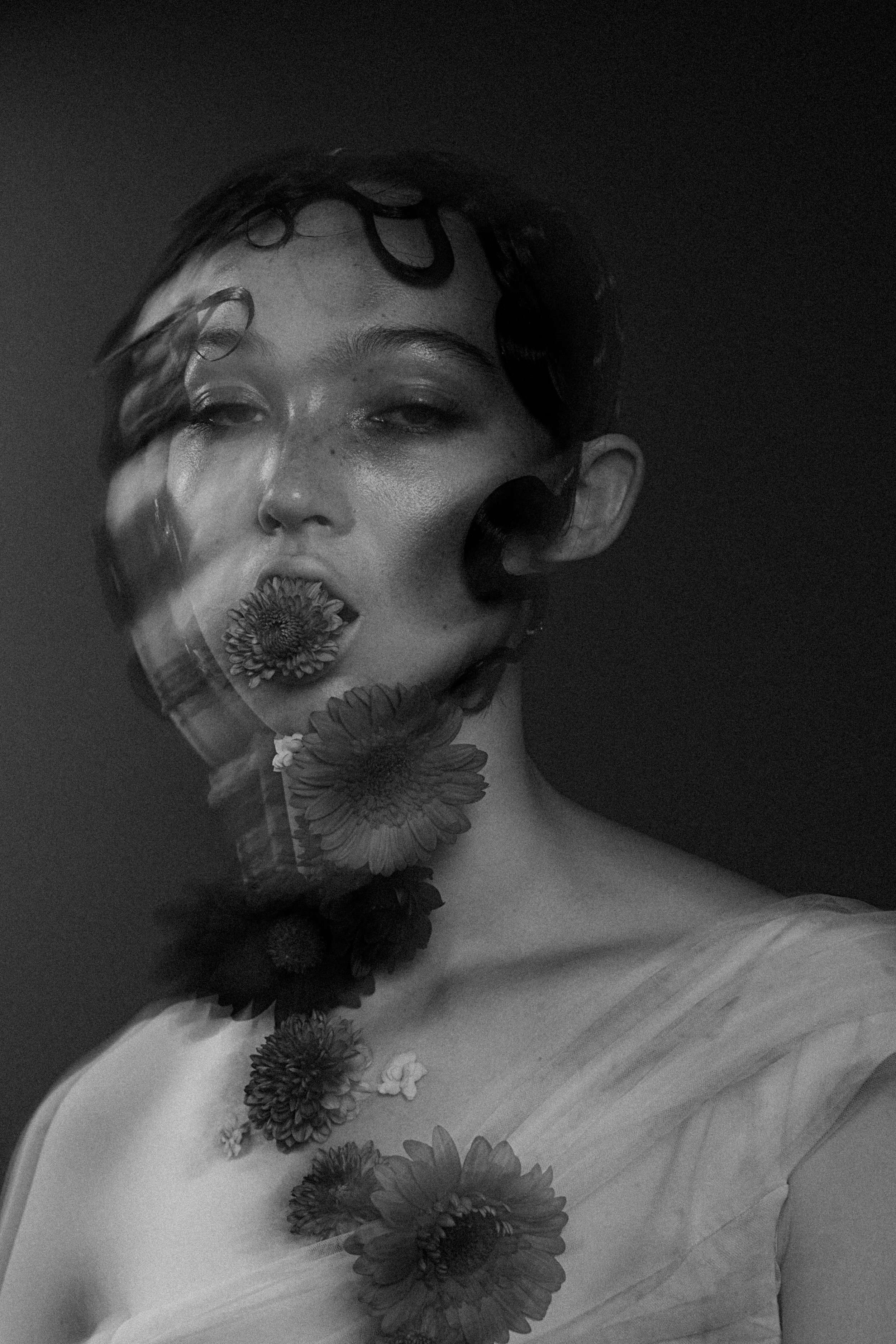 A black and white portrait of a woman with short hair, flowers around her face and on her shoulder, with her eyes half-closed, wearing a soft, sheer fabric on her shoulder.