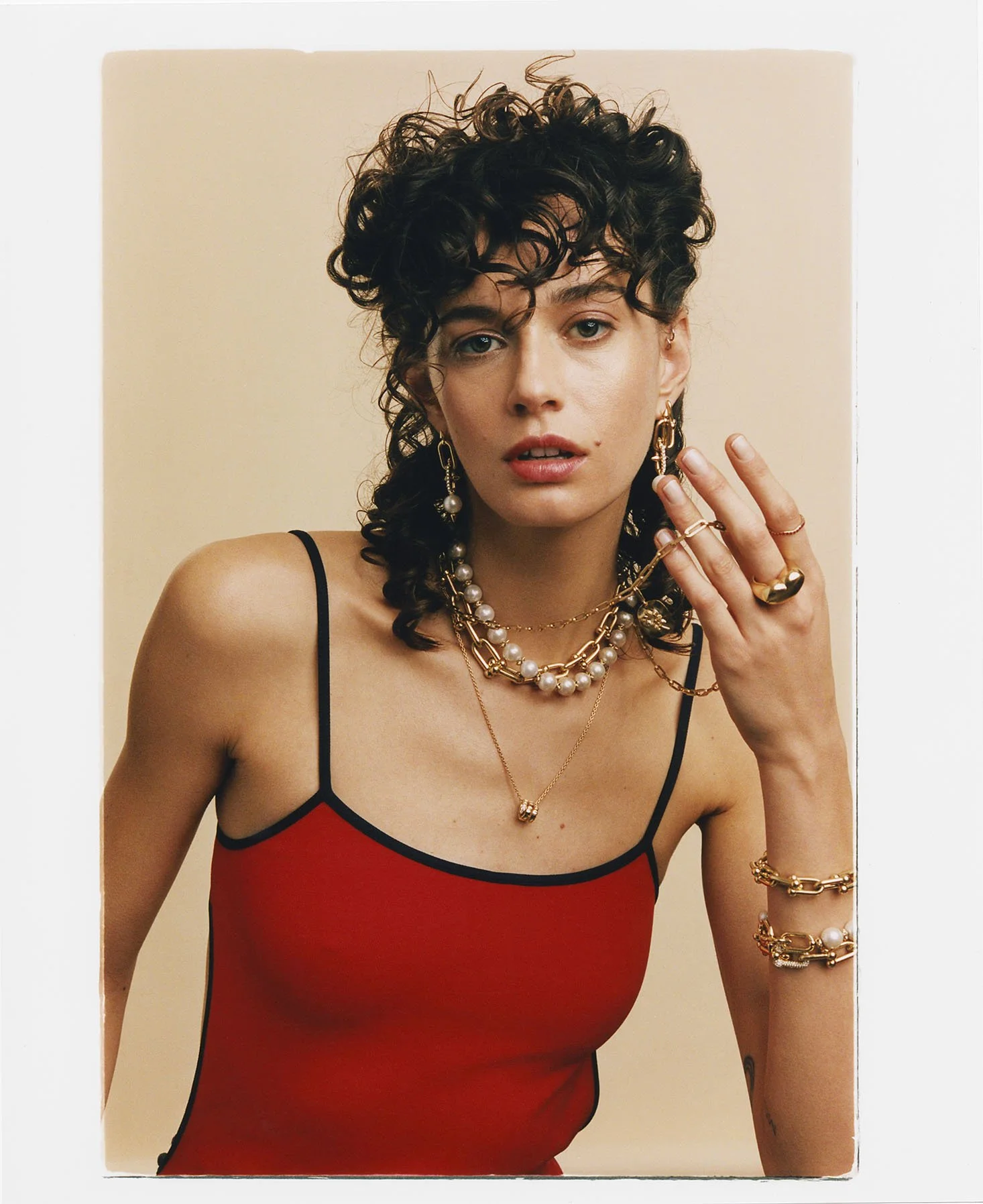 A woman with curly hair wearing a red top with black straps, adorned with gold and pearl jewelry including necklaces, earrings, bracelets, and rings.