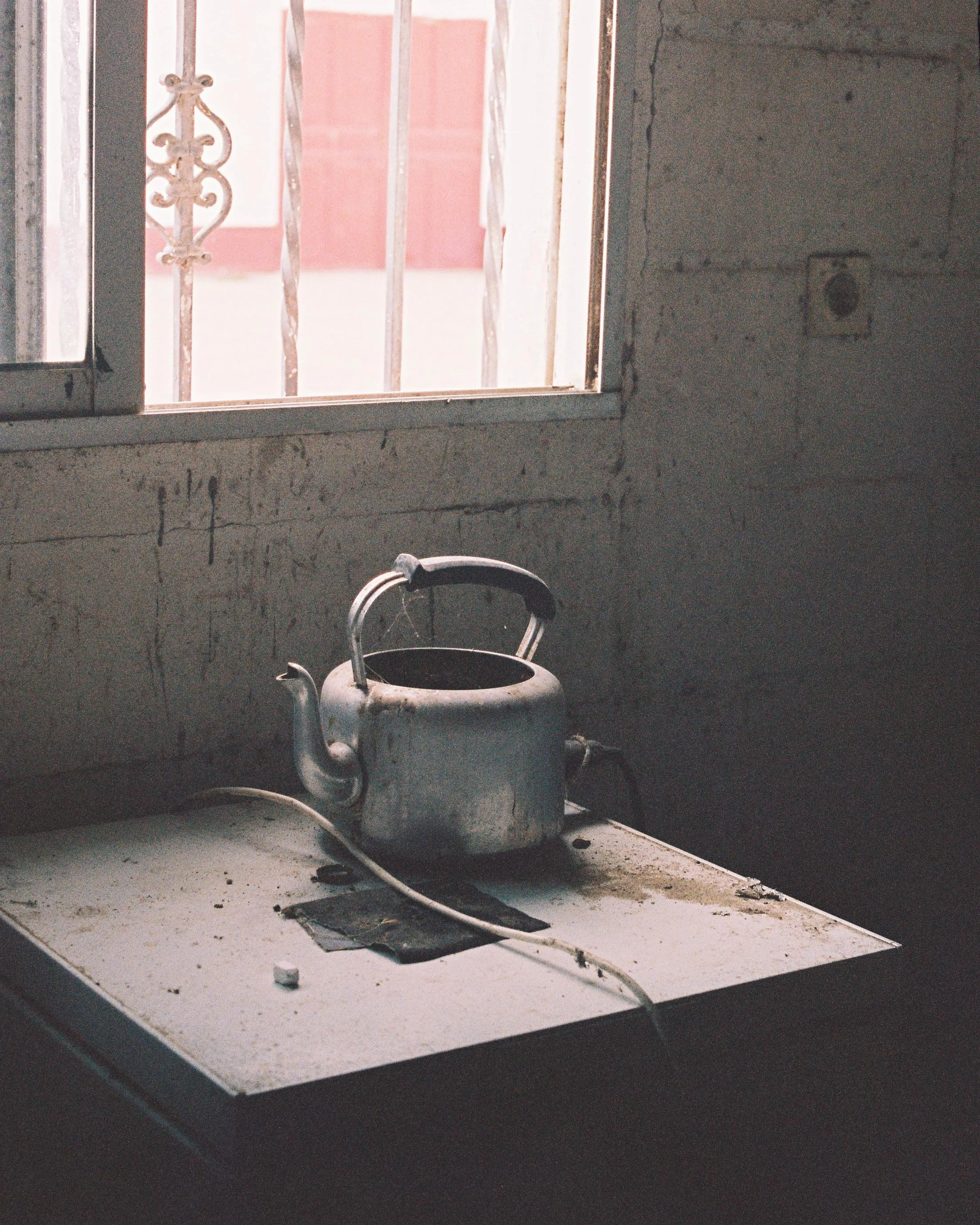 Old, dusty kettle on a worn white stove with a window in the background, exposing rusty metal bars and a pink wall outside.