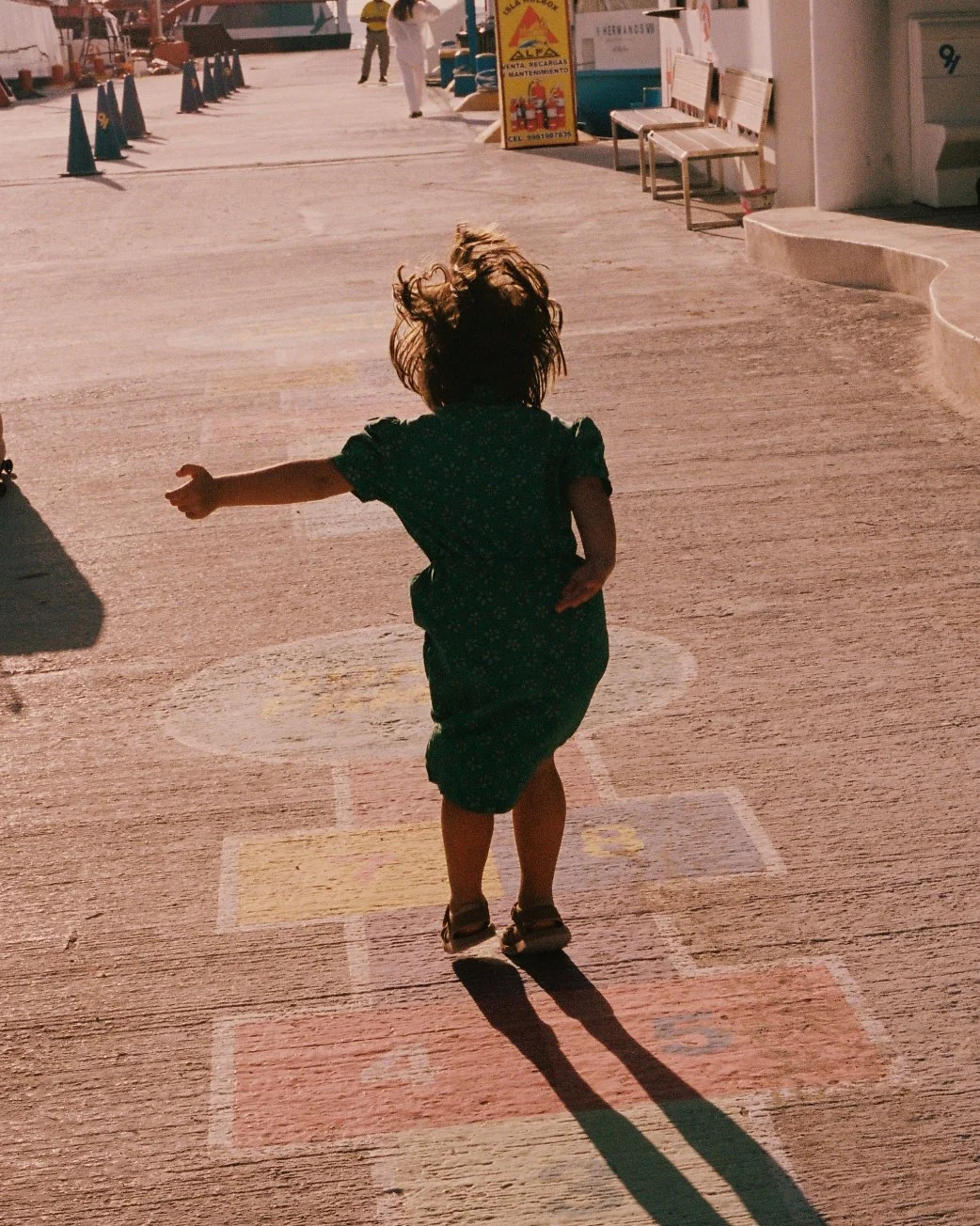 Child playing on a hopscotch game drawn on the pavement, with a shadow cast behind them, in an outdoor area with benches and signs.