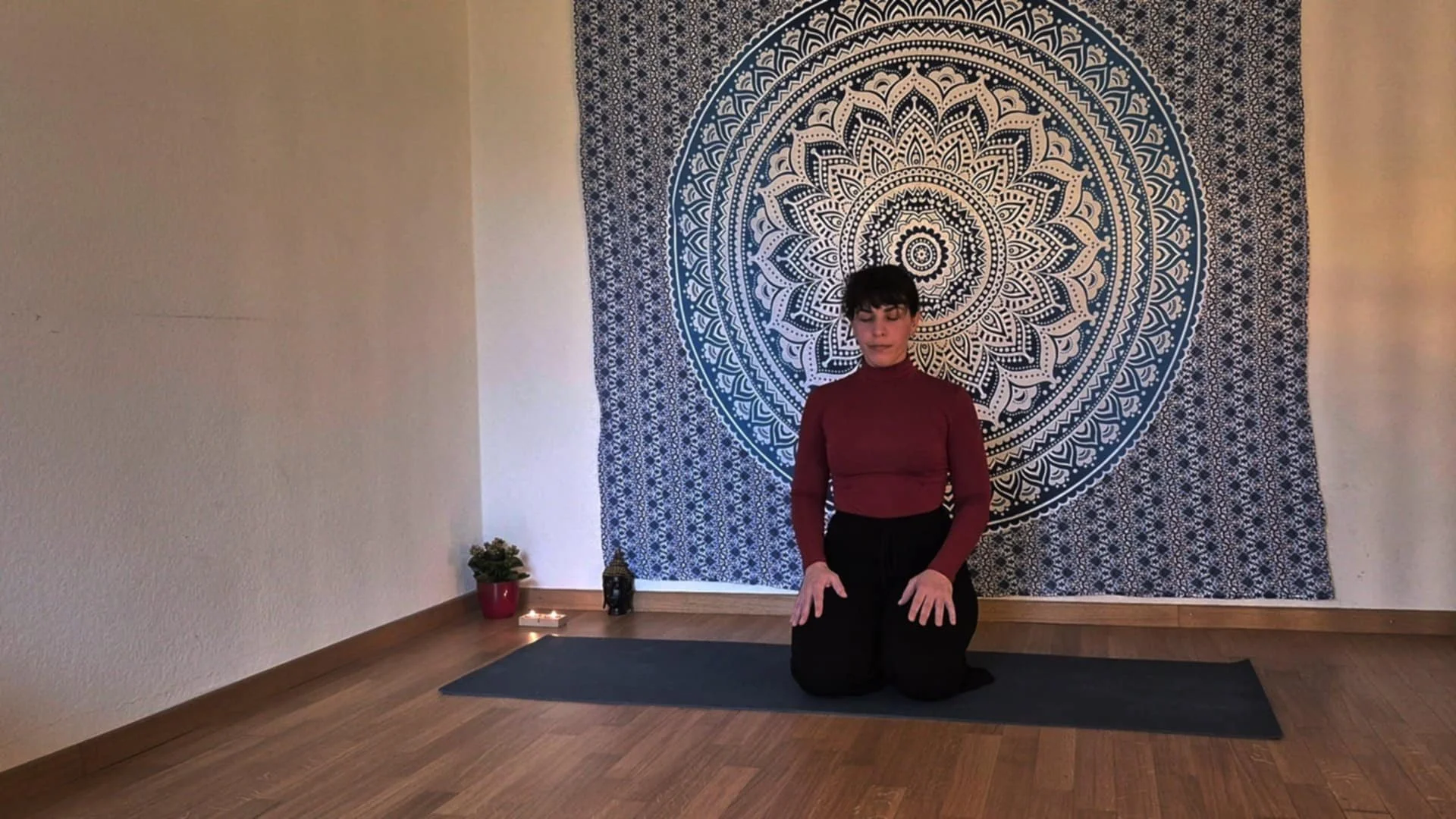 Woman practicing yoga on a mat with a mandala tapestry and candle in a peaceful room.