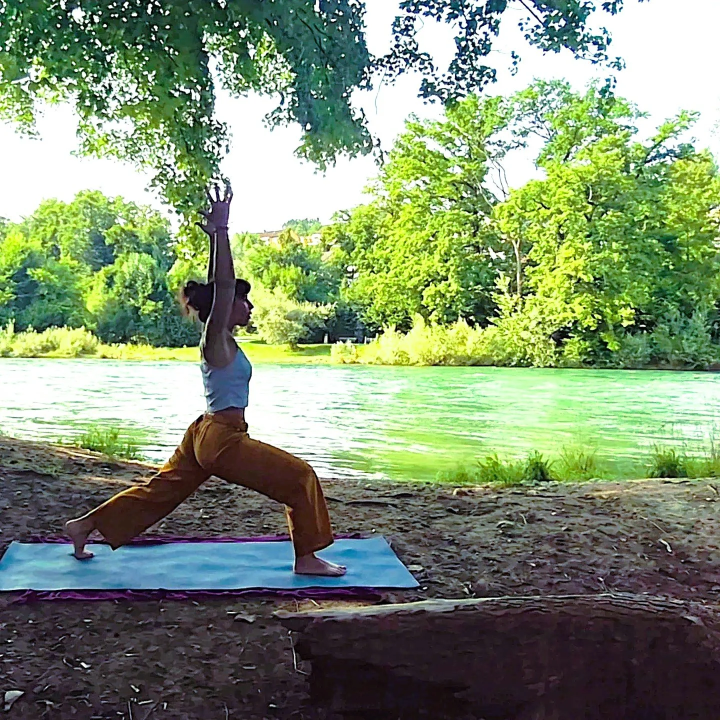 A woman practicing yoga in a warrior pose outdoors on a yoga mat by a river, surrounded by lush green trees and clear weather.