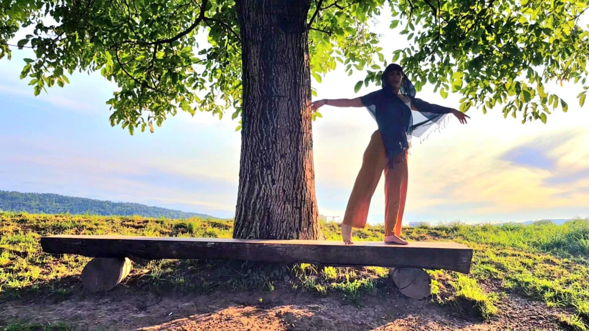 A woman standing on a wooden plank in front of a large tree, with her arms outstretched, outdoors during sunset, with a landscape of grass, hills, and a partly cloudy sky in the background.