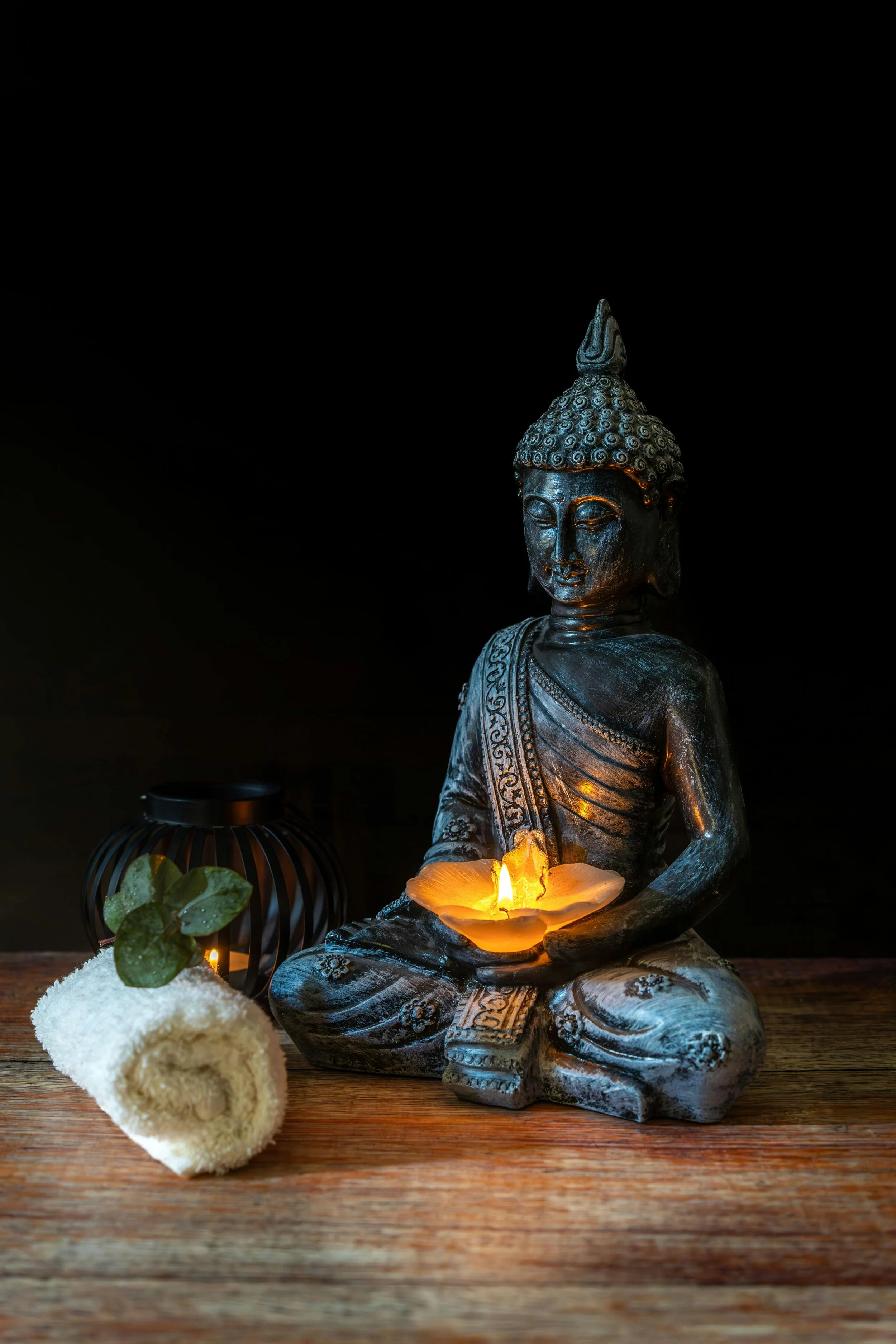 Buddha statue holding a candle, surrounded by a rolled towel with leaves, a lit candle, and a decorative black lantern on a wooden surface, against a dark background.