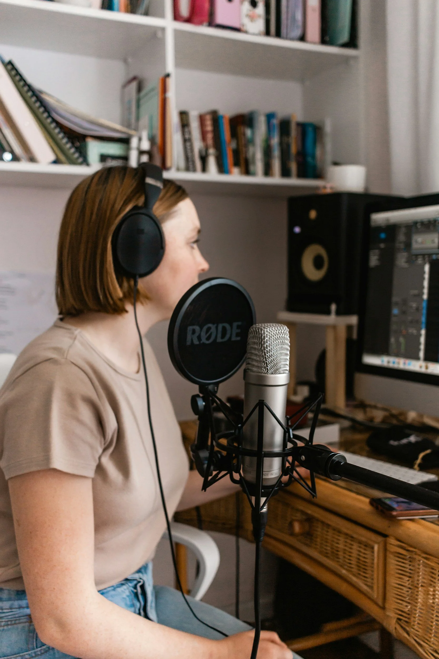 Eine junge Frau mit Short und T-Shirt trägt Kopfhörer und singt in ein Mikrofon in einem Raum mit einem Regal voller Bücher, einem Computer und einem Lautsprecher.