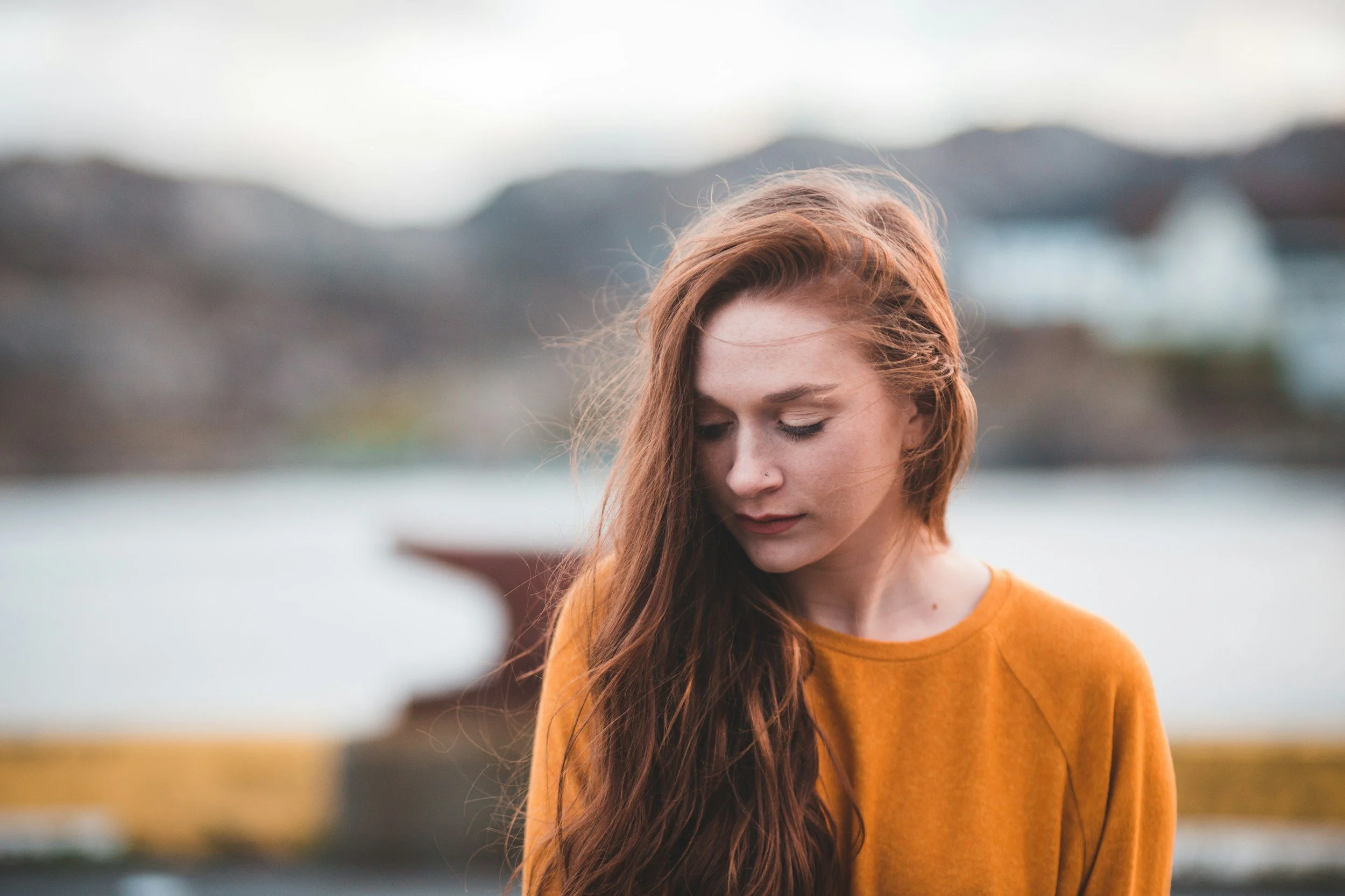 Junge Frau mit langen roten Haaren, die ihre Augen schließt, vor unscharfer Wasser- und Bergkulisse, trägt ein orangefarbenes Oberteil.