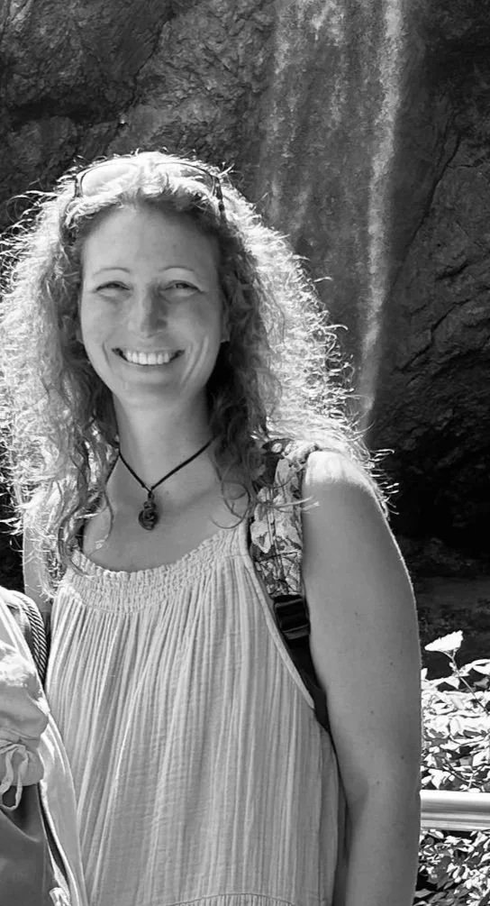 Eine lächelnde Frau mit Locken, trägt ein ärmelloses, gestricktes Oberteil, mit einem Rucksack und einem Halsband, im Freien vor einer Felswand.