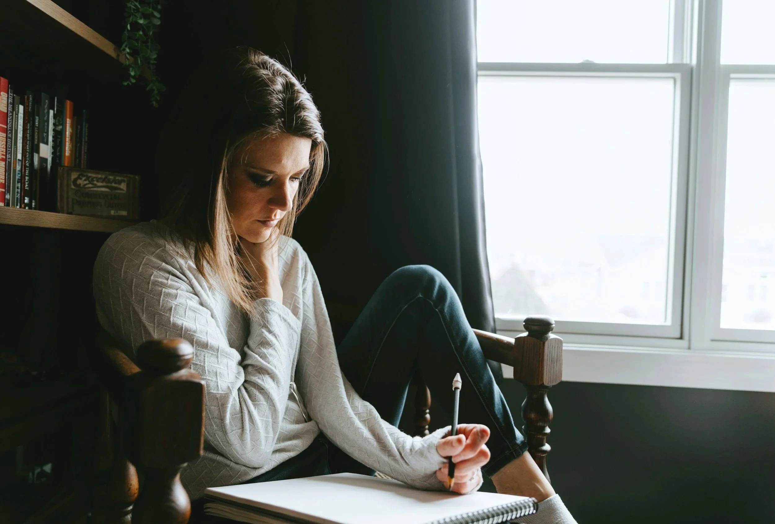 Eine junge Frau sitzt in einem dunklen Raum neben einem Fenster, liest oder schreibt in ein Notebook, mit einem Notizbuch vor sich. Sie trägt einen weißen Pullover, ihre Haare sind braun und offen. Im Hintergrund sind Bücherregale sichtbar.
