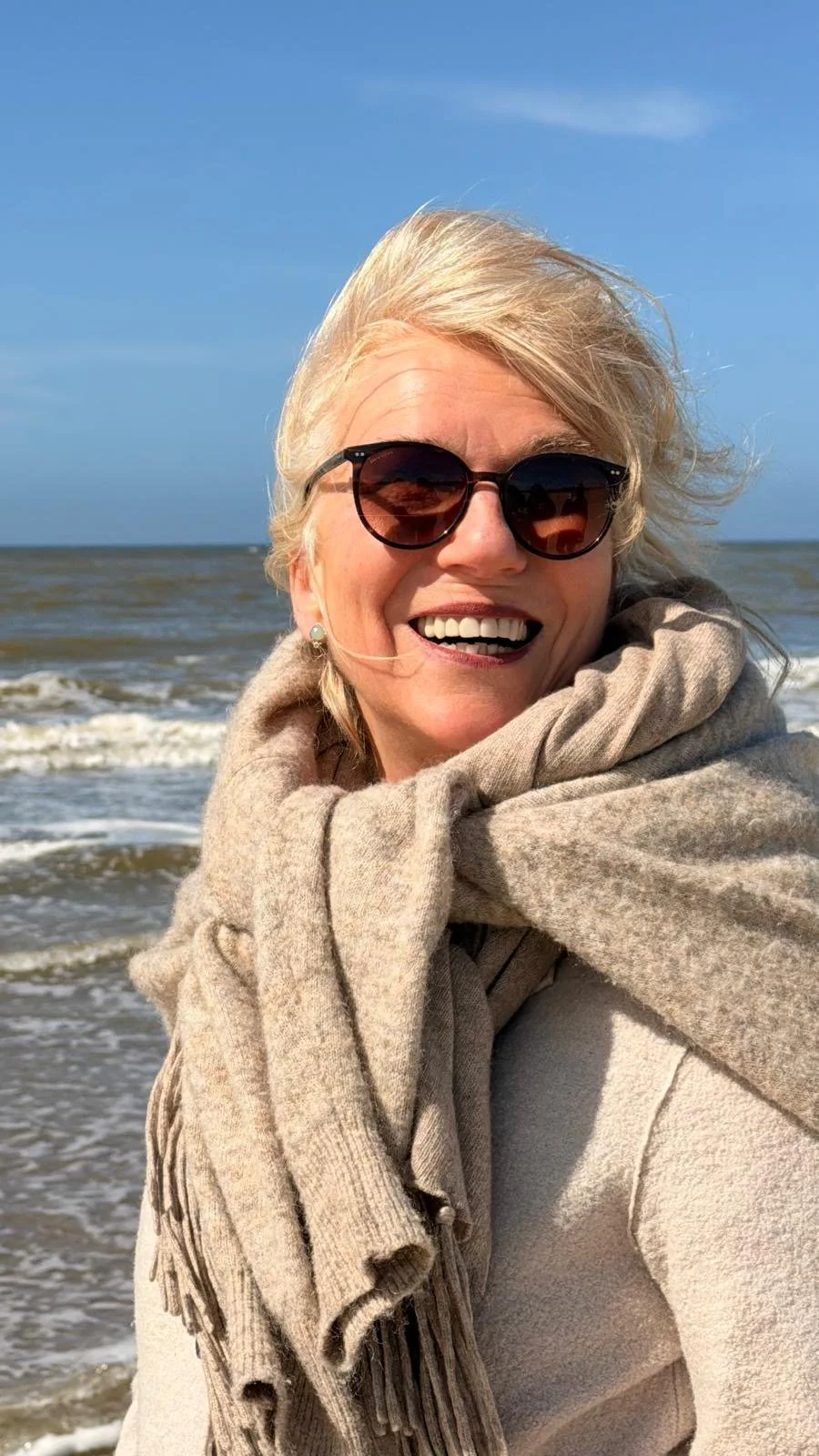 Lächelnde ältere Frau mit dunklen Sonnenbrillen, grauem Schal und cremefarbenem Mantel am Strand bei Sonne, Meer im Hintergrund.