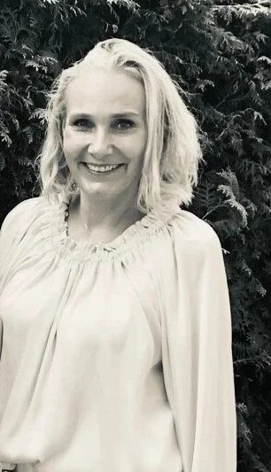Eine lächelnde Frau mit blonden Haaren vor einem dunklen, bewaldeten Hintergrund, in Schwarz-Weiß