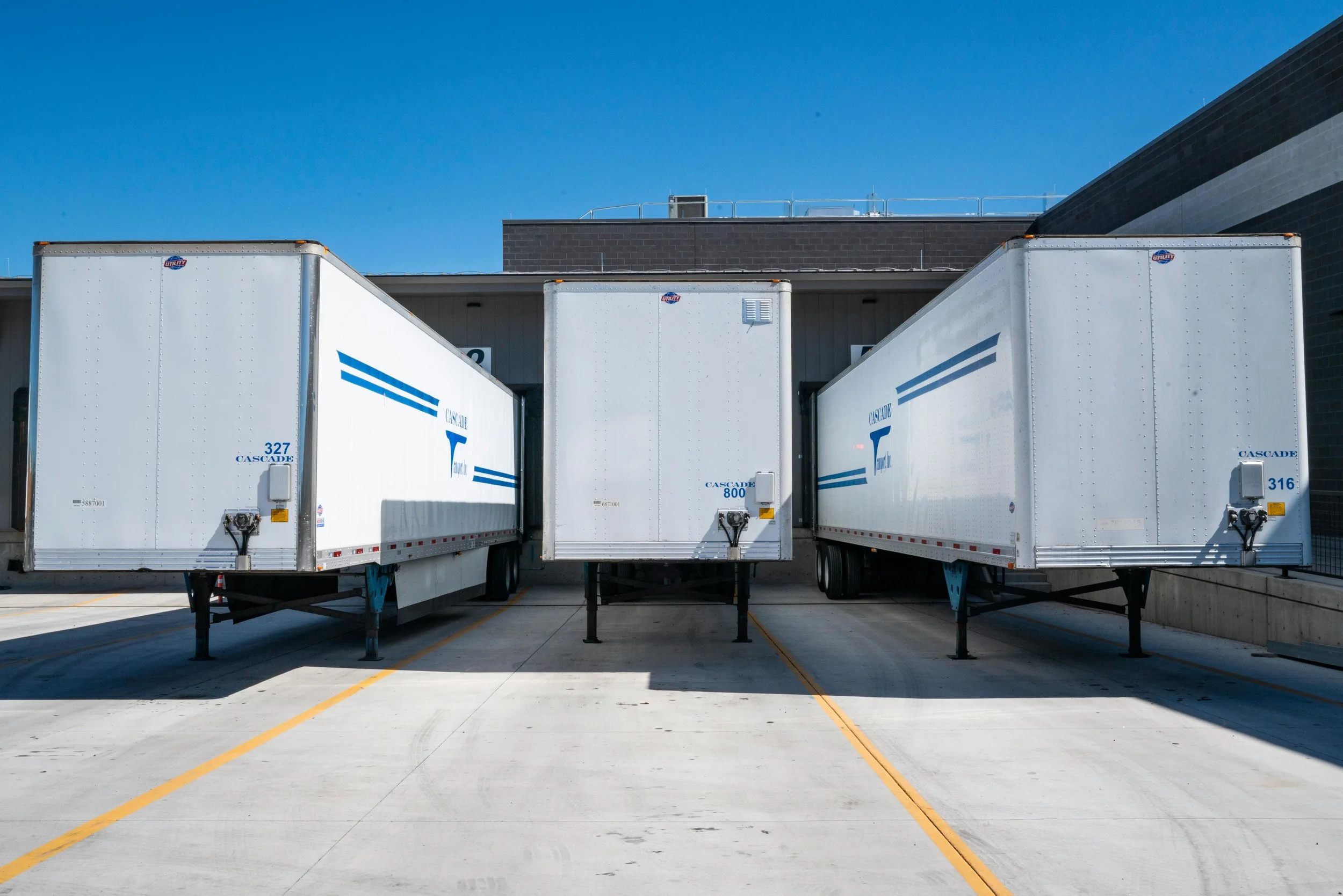 Three semi-trailers parked side by side in a loading dock area under a clear blue sky.