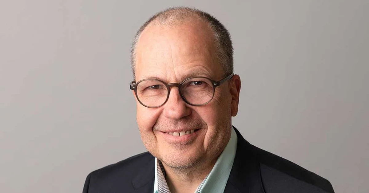 A professional headshot of a middle-aged man with glasses, smiling, wearing a dark suit jacket and a collared shirt, against a plain background.