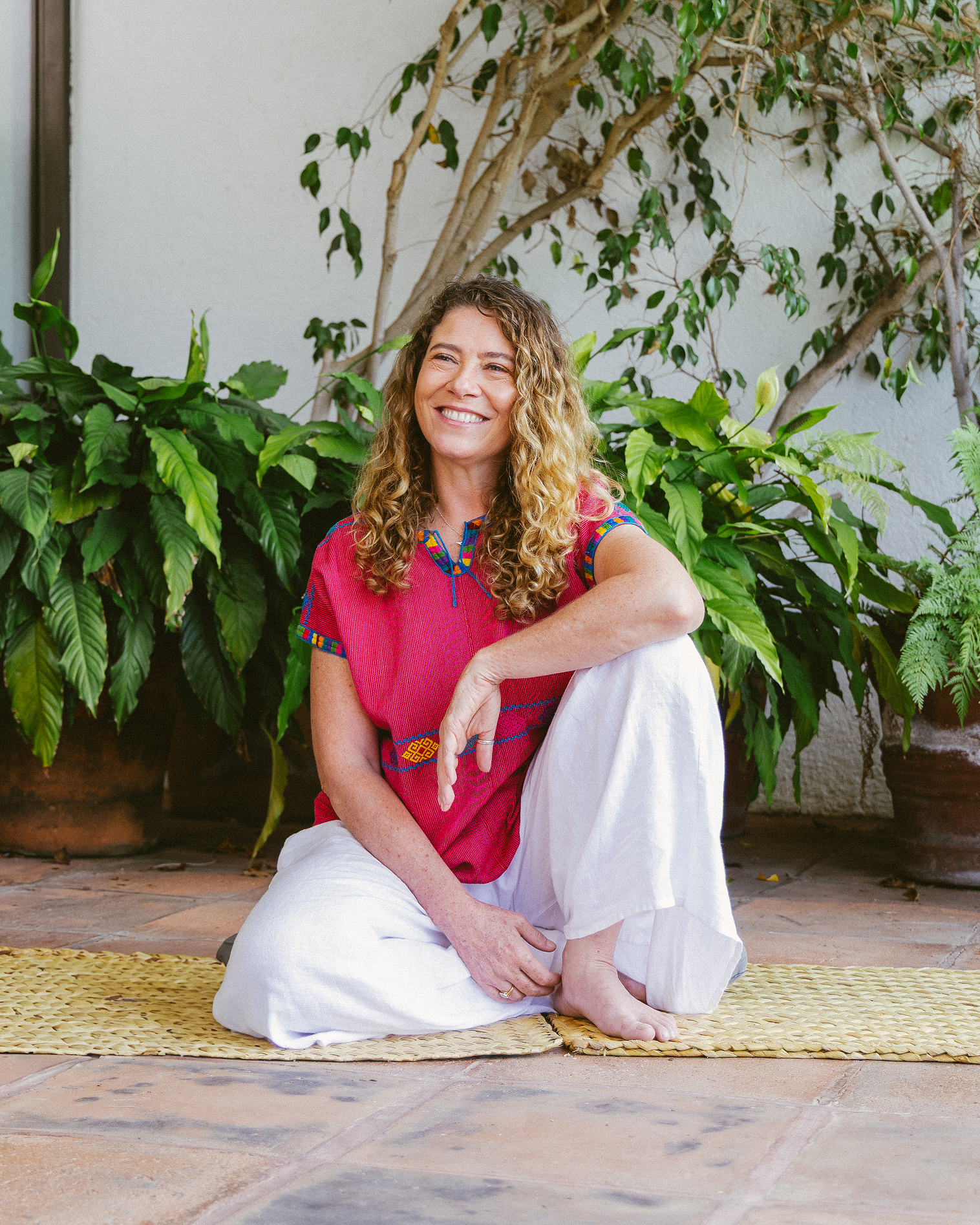 A woman sitting on a yellow mat outdoors, smiling, with large green plants and trees in the background, wearing a red top and white pants.