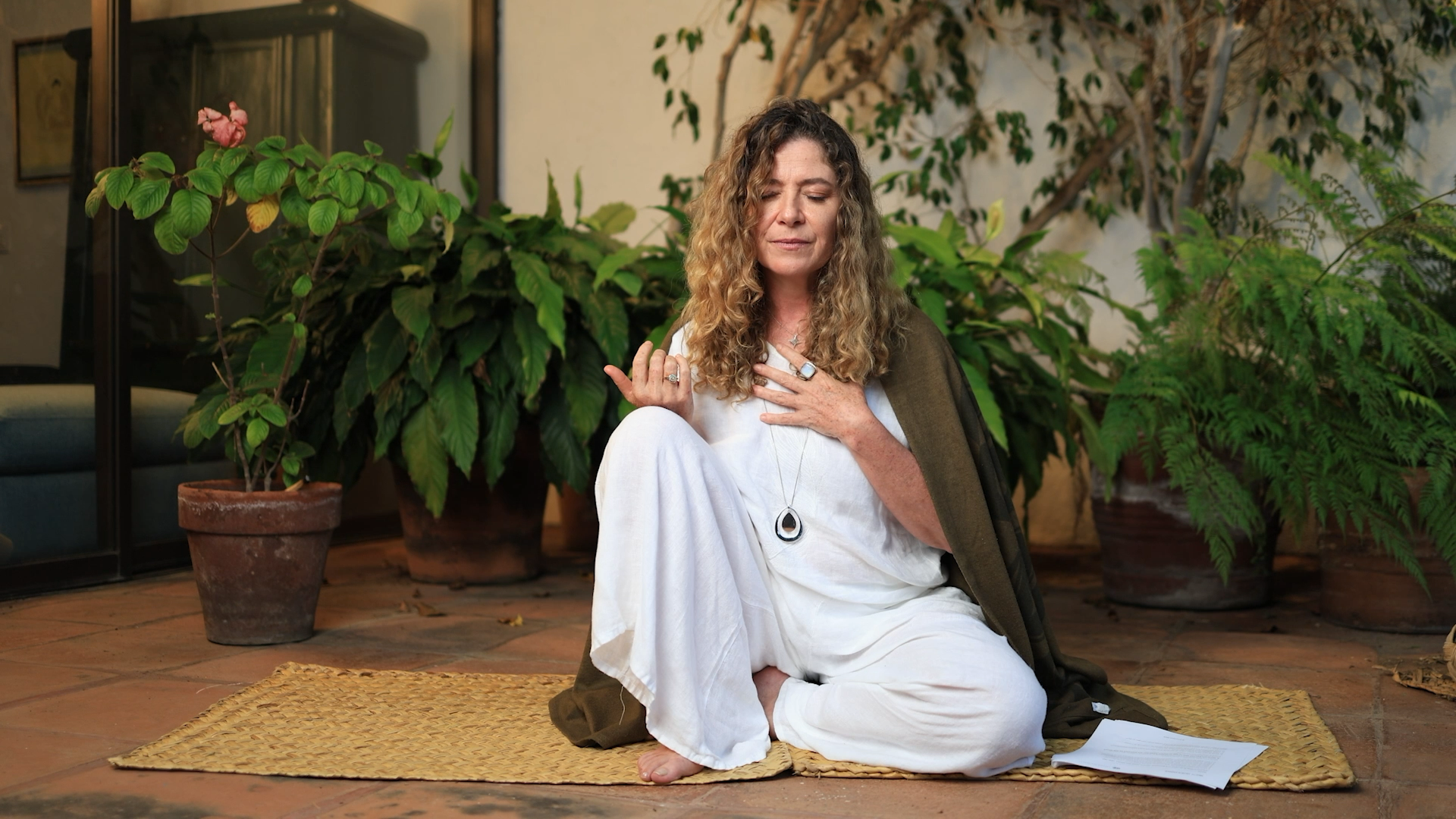 A woman with curly hair sitting cross-legged on a woven mat on a tile floor, surrounded by potted plants, with her eyes closed and one hand on her chest, holding a piece of paper nearby.