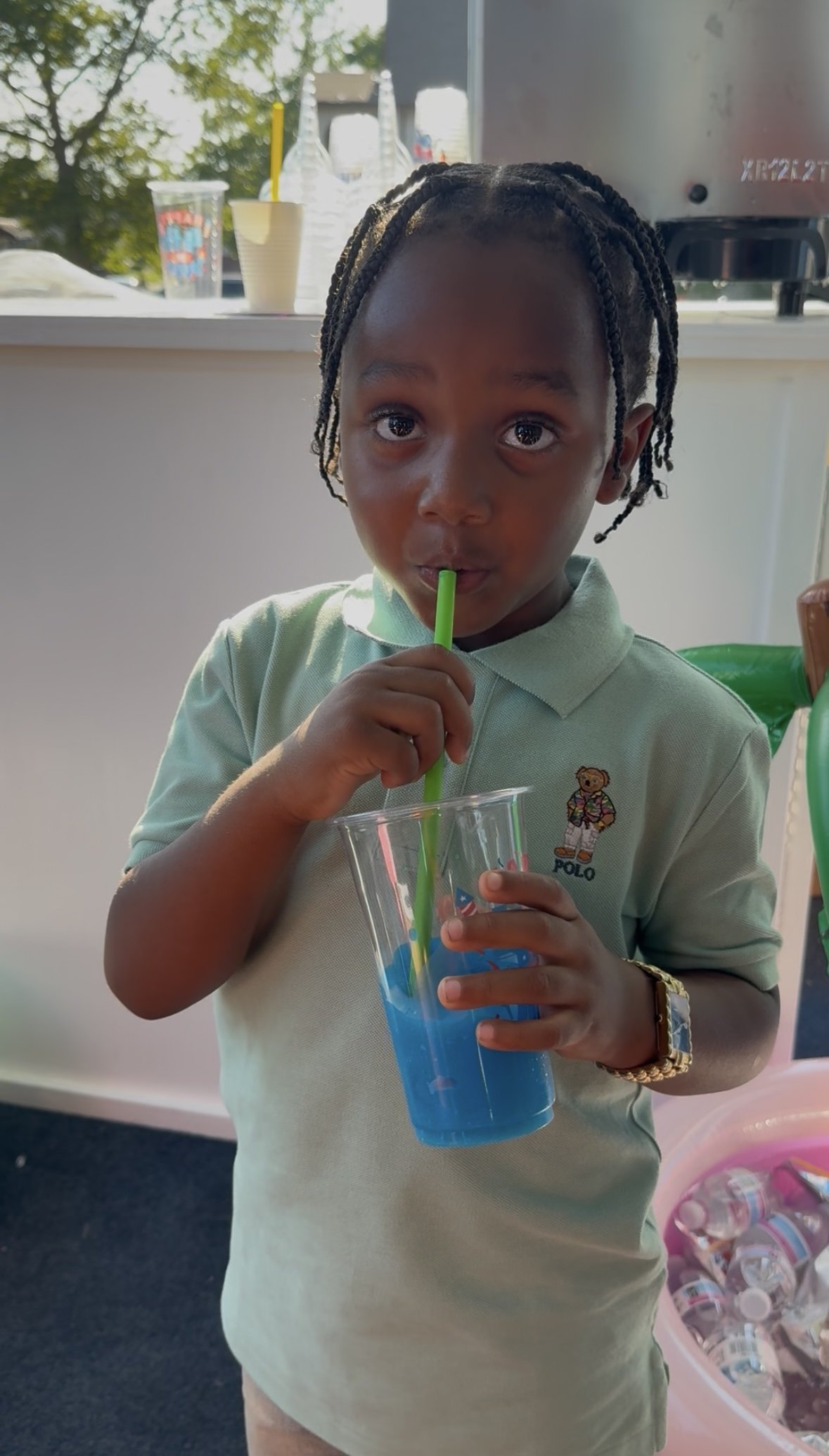 A young boy with braided hair sipping blue drink through a bright green straw, wearing a light green polo shirt with a teddy bear logo, standing indoors near a counter with bottles and cups in the background.