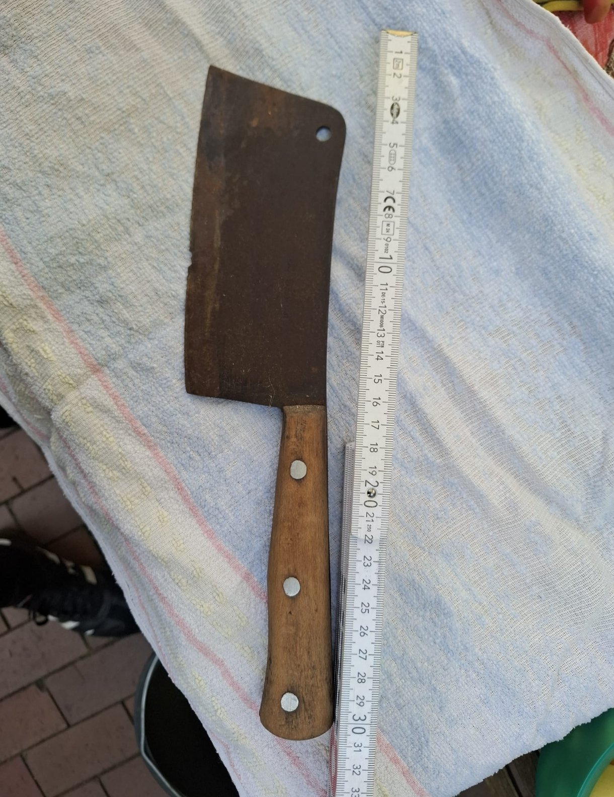 Ein alter, rustikaler Hackebeil mit Holzgriff auf einem bunten Stofftuch neben einem Maßlineal.