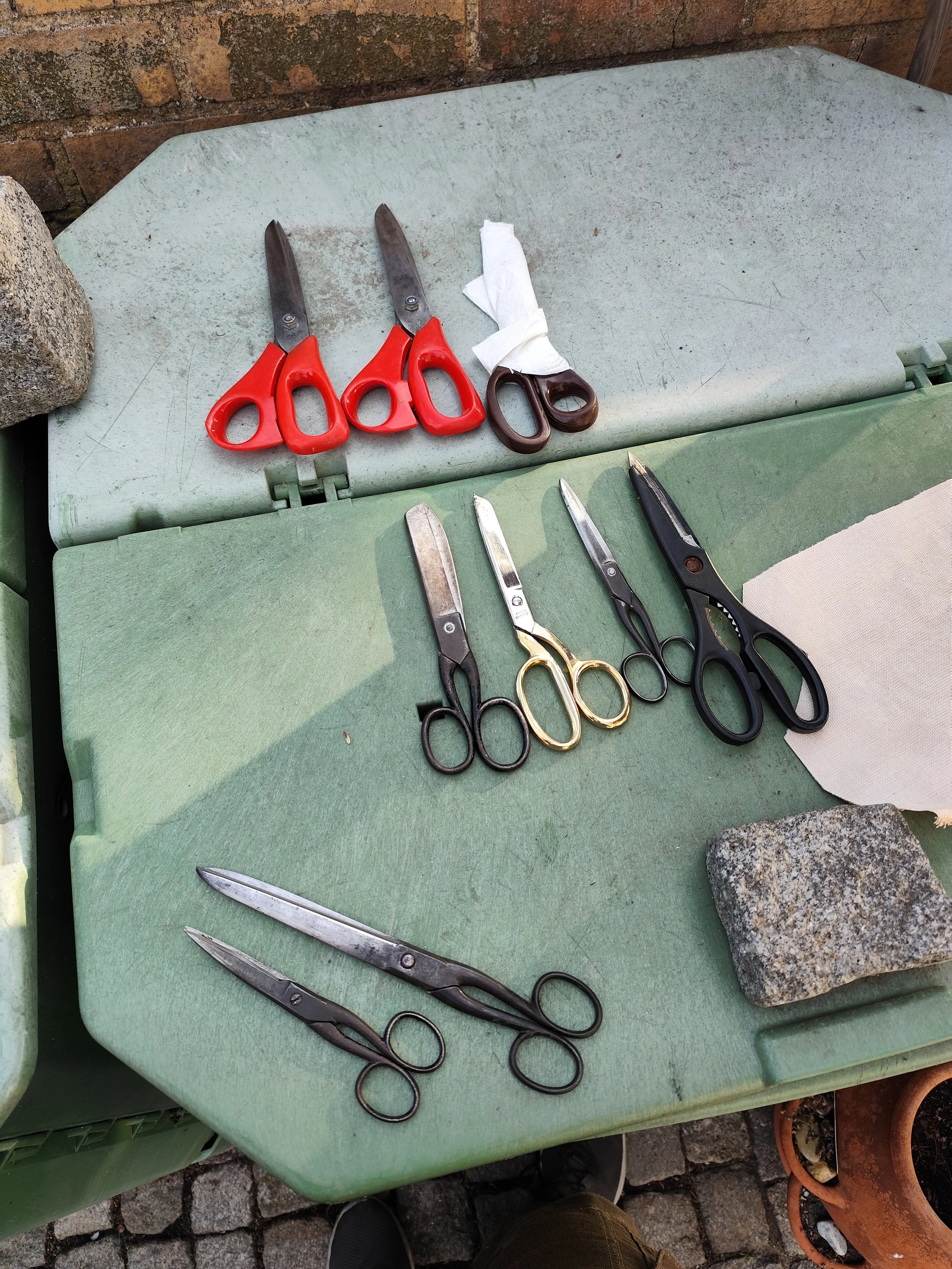 Verschiedene Gartenscheren und Gartengeräte auf einer grünen Arbeitsfläche, umgeben von Steinen und einer Mauer.