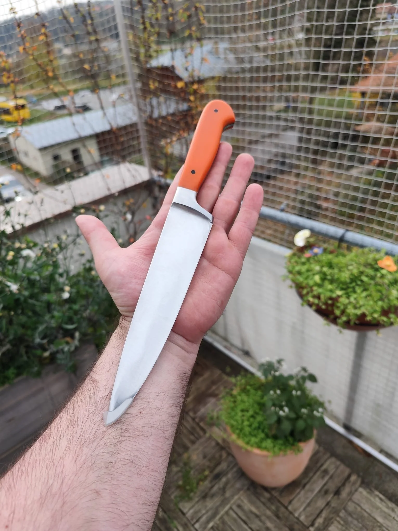 Hand hält ein großes Küchenmesser mit orangeem Griff auf einem Balkon mit Pflanzen im Hintergrund.