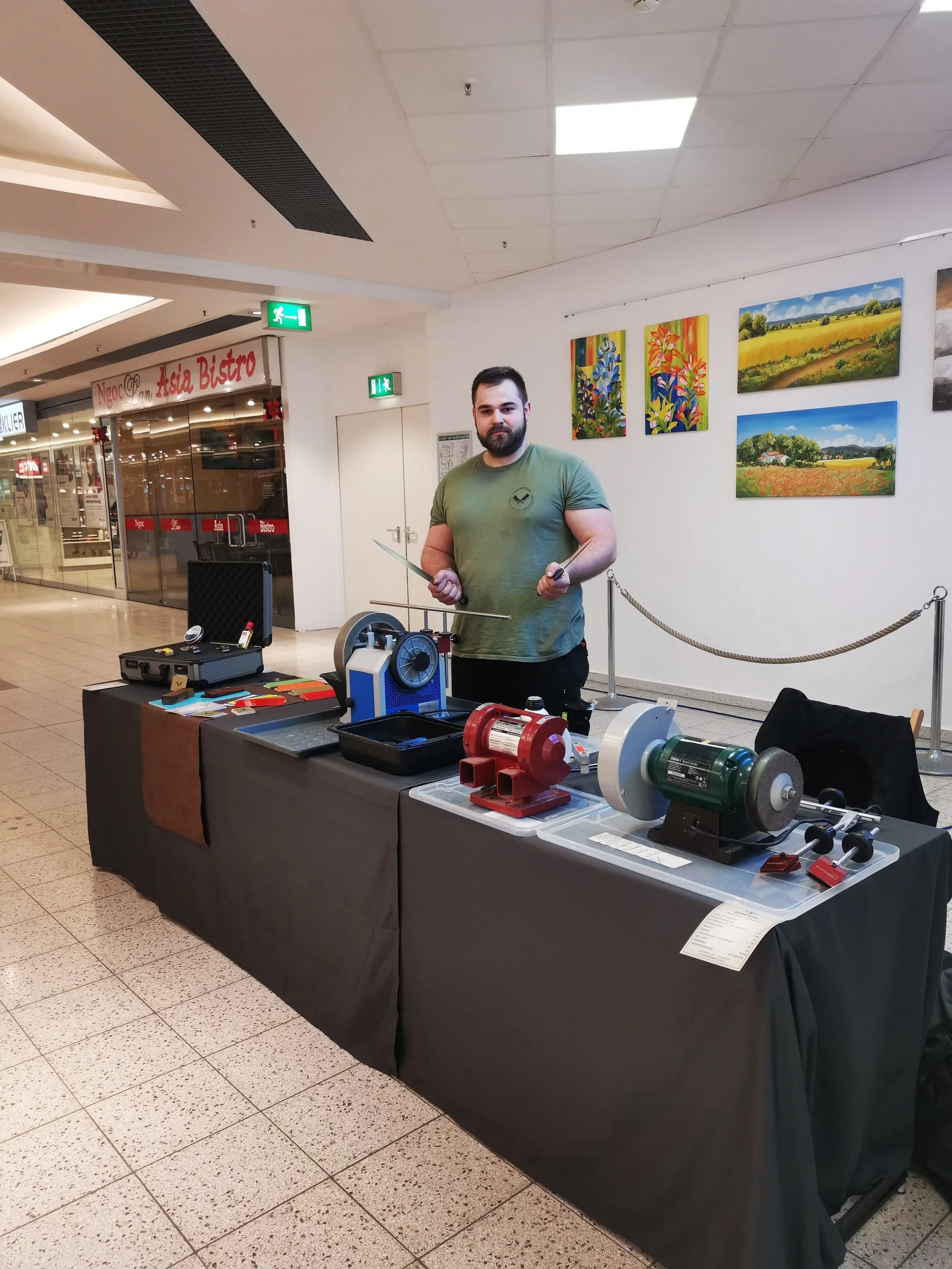 Ein Mann steht hinter einem Tisch mit verschiedenen Maschinen und Werkzeugen, in einem Innenraum mit Kunstwerken an der Wand.