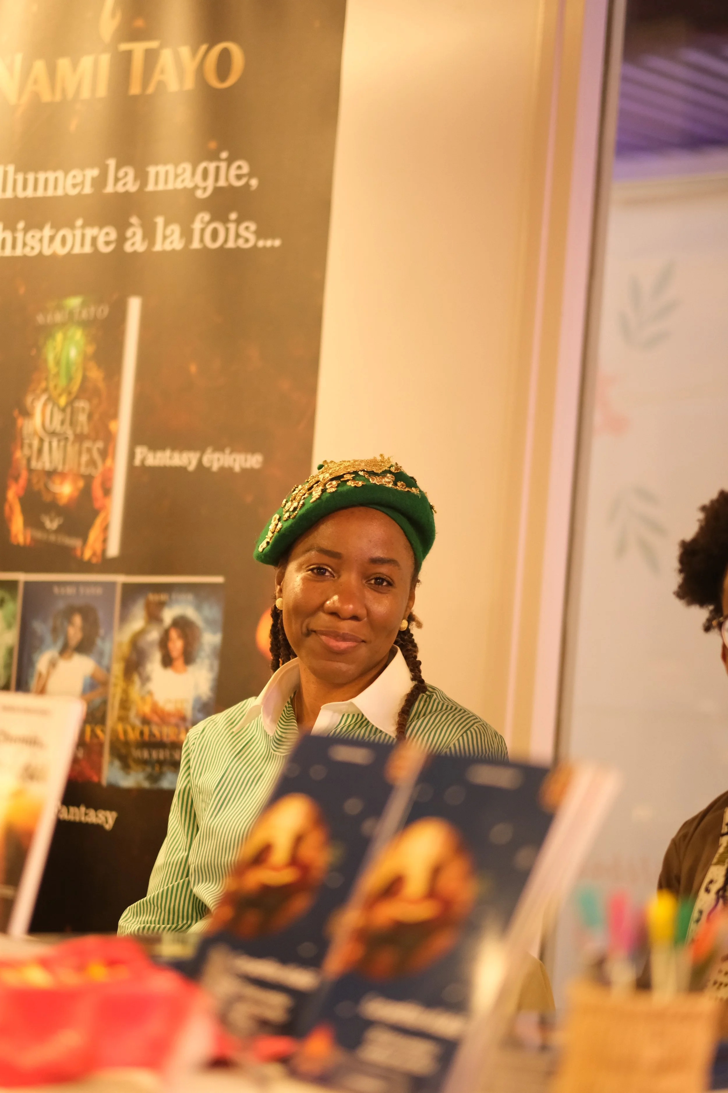 Une femme souriante portant un couvre-chef vert avec des ornements dorés, debout à un stand avec des affiches promotionnelles de films ou de séries en arrière-plan.