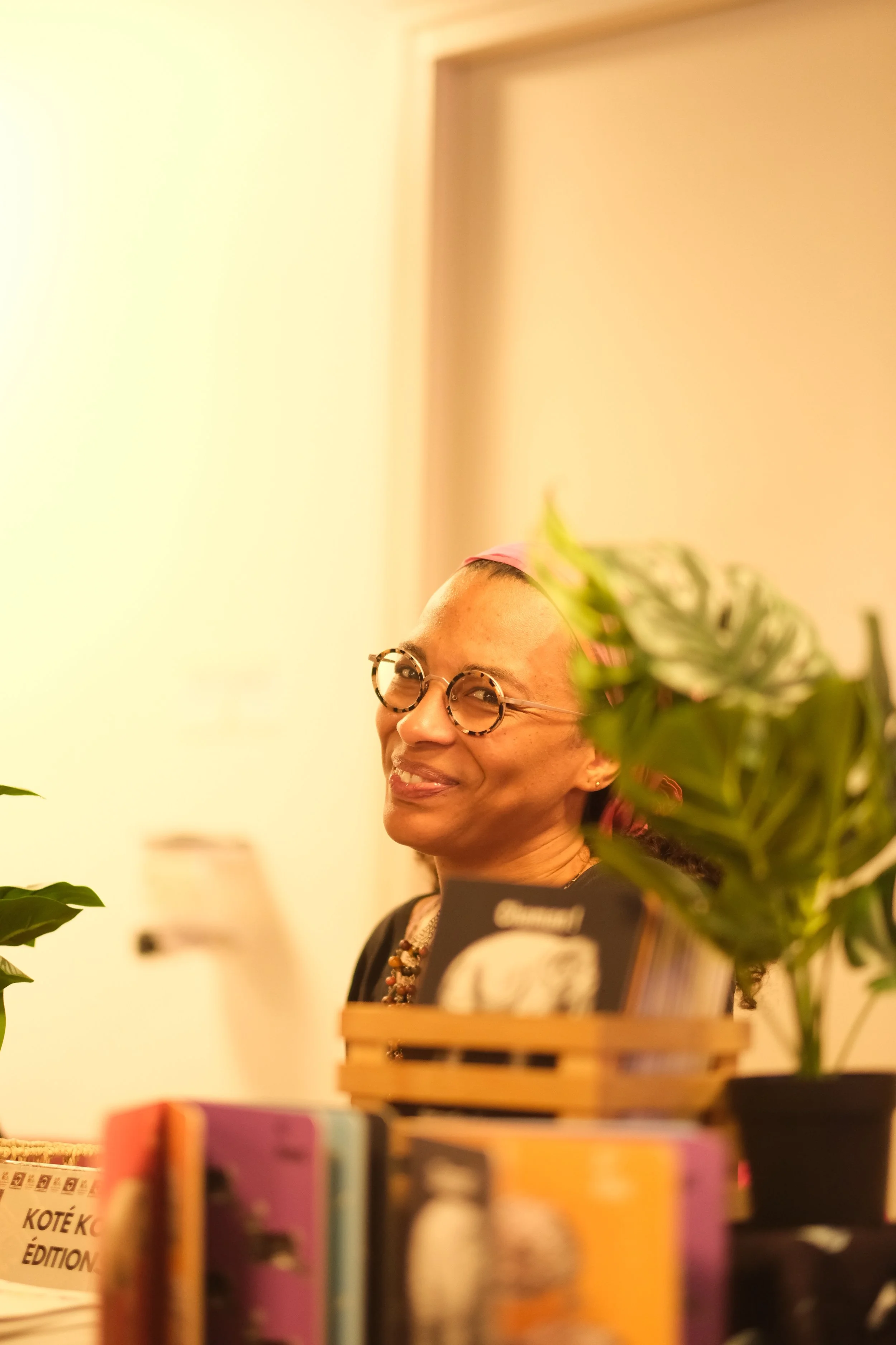 Une femme souriante portant des lunettes, entourée de livres et de plantes dans un environnement intérieur chaleureux.