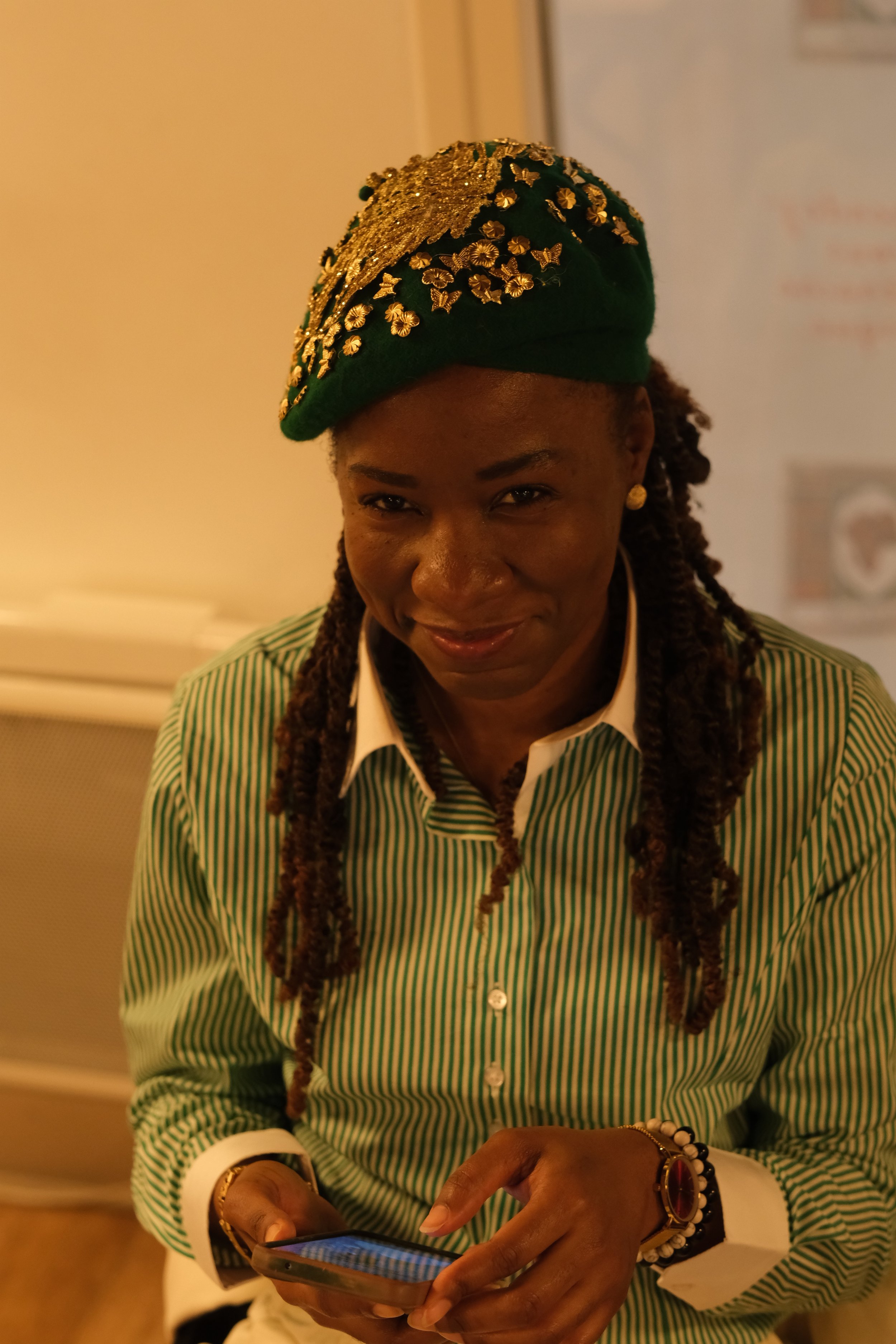 Une femme porte un béret vert orné de motifs dorés et une chemise à rayures vertes et blanches. Elle regarde un téléphone portable et sourit légèrement.