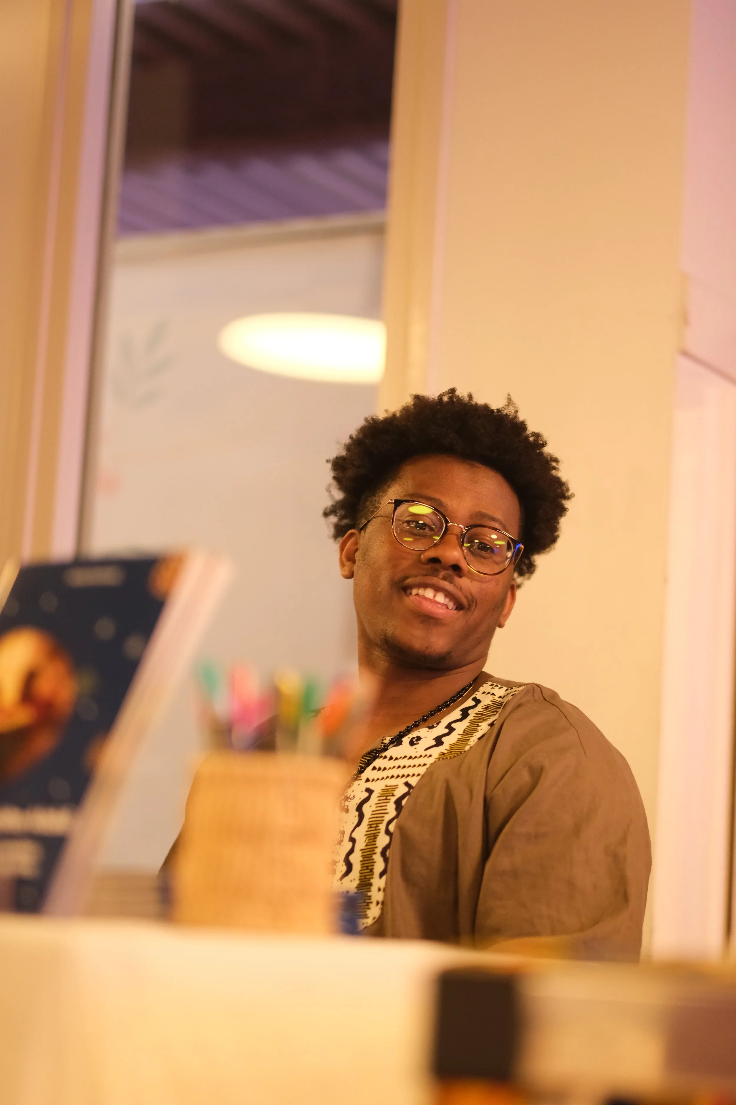 Un jeune homme souriant portant des lunettes, assis à une table dans un intérieur avec des murs de couleur beige et un plafond en bois, prenant une photo de lui-même avec un téléphone.