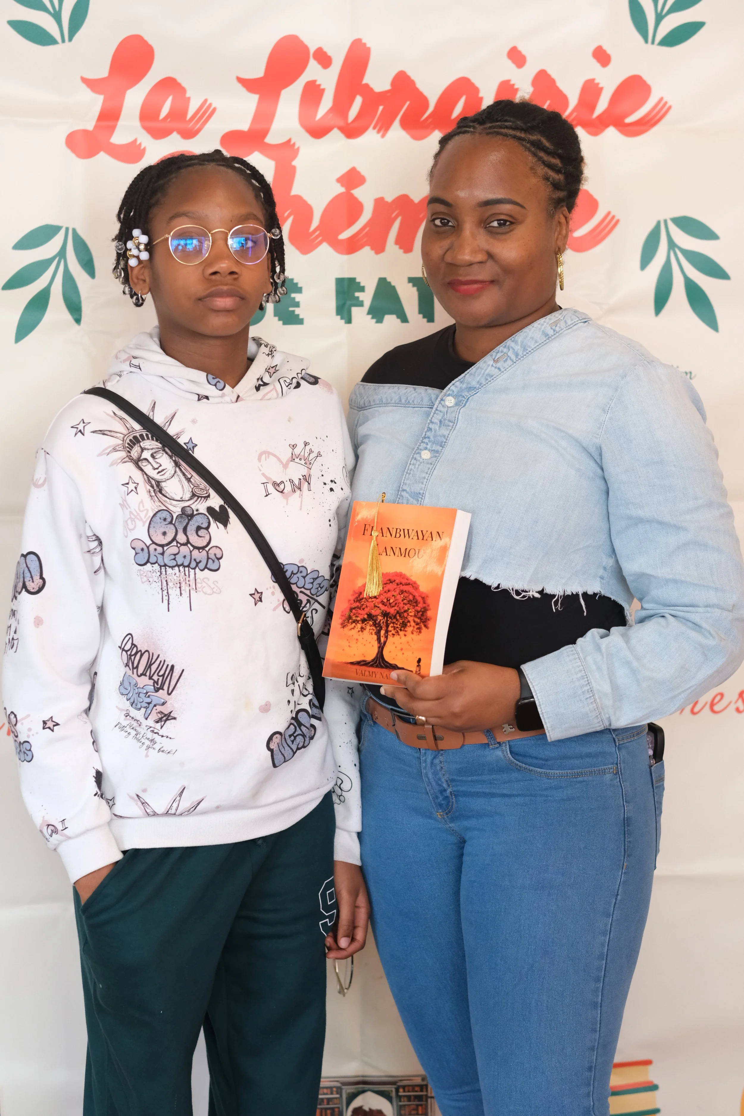 Deux femmes posent ensemble lors d'un événement, derrière elles un panneau avec le texte 'La librairie' et des illustrations de feuilles.