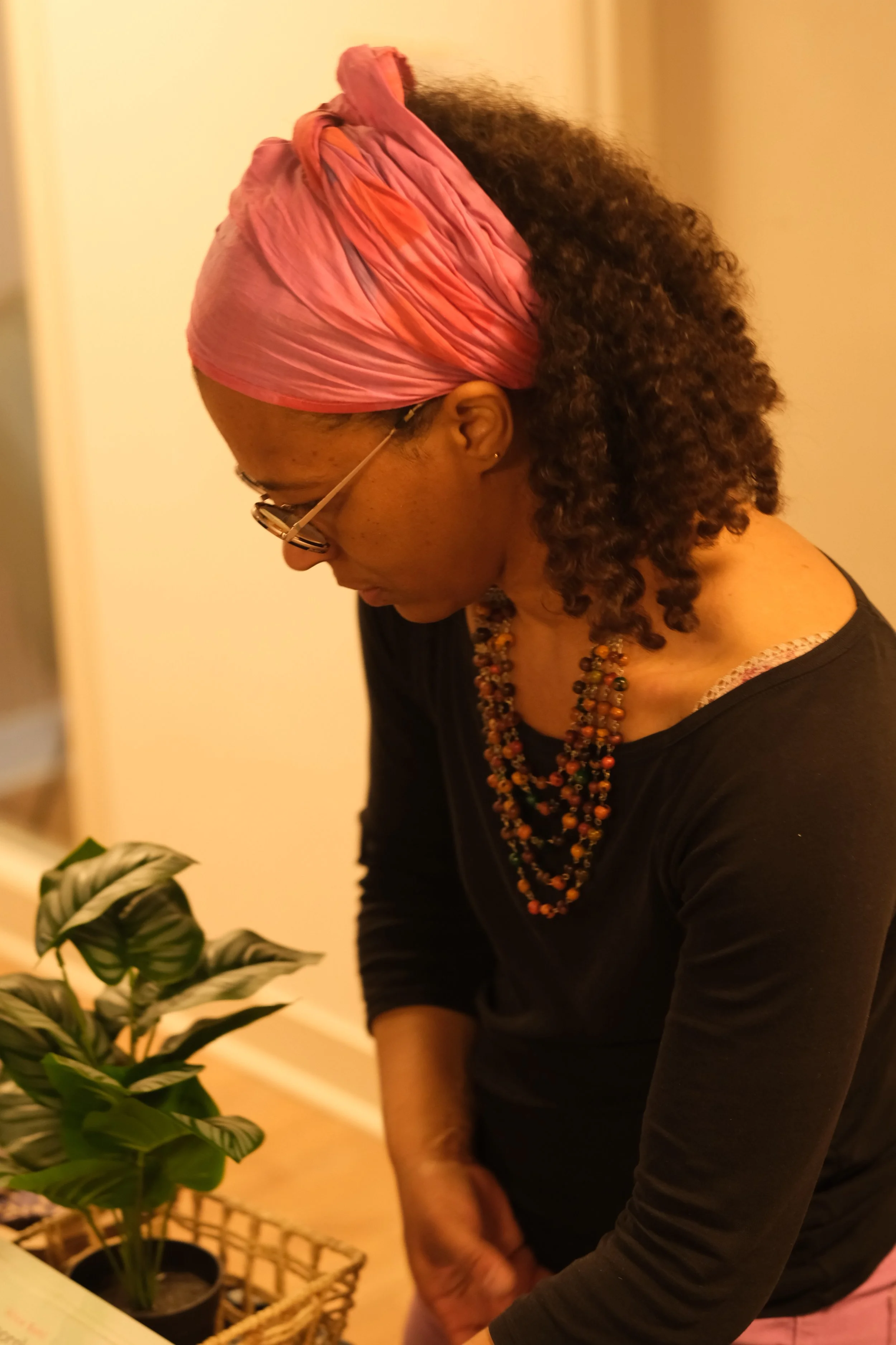 Une femme portant un foulard rose, lunettes et collier coloré, penchée en avant près d'une plante verte en pot.