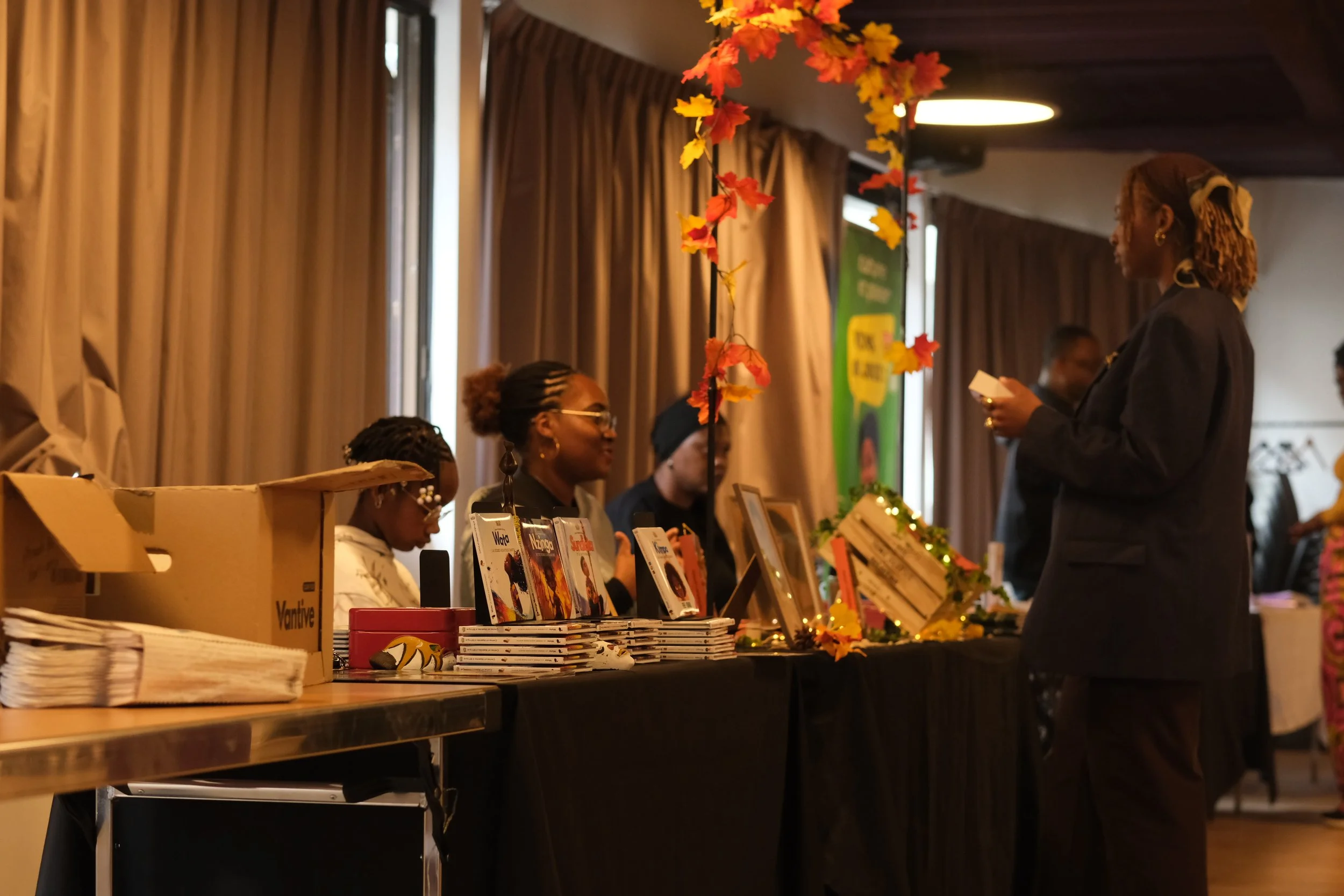 Une femme parle à un stand de livres lors d'un événement intérieur en salle, décoré de feuilles d'automne en plastique.