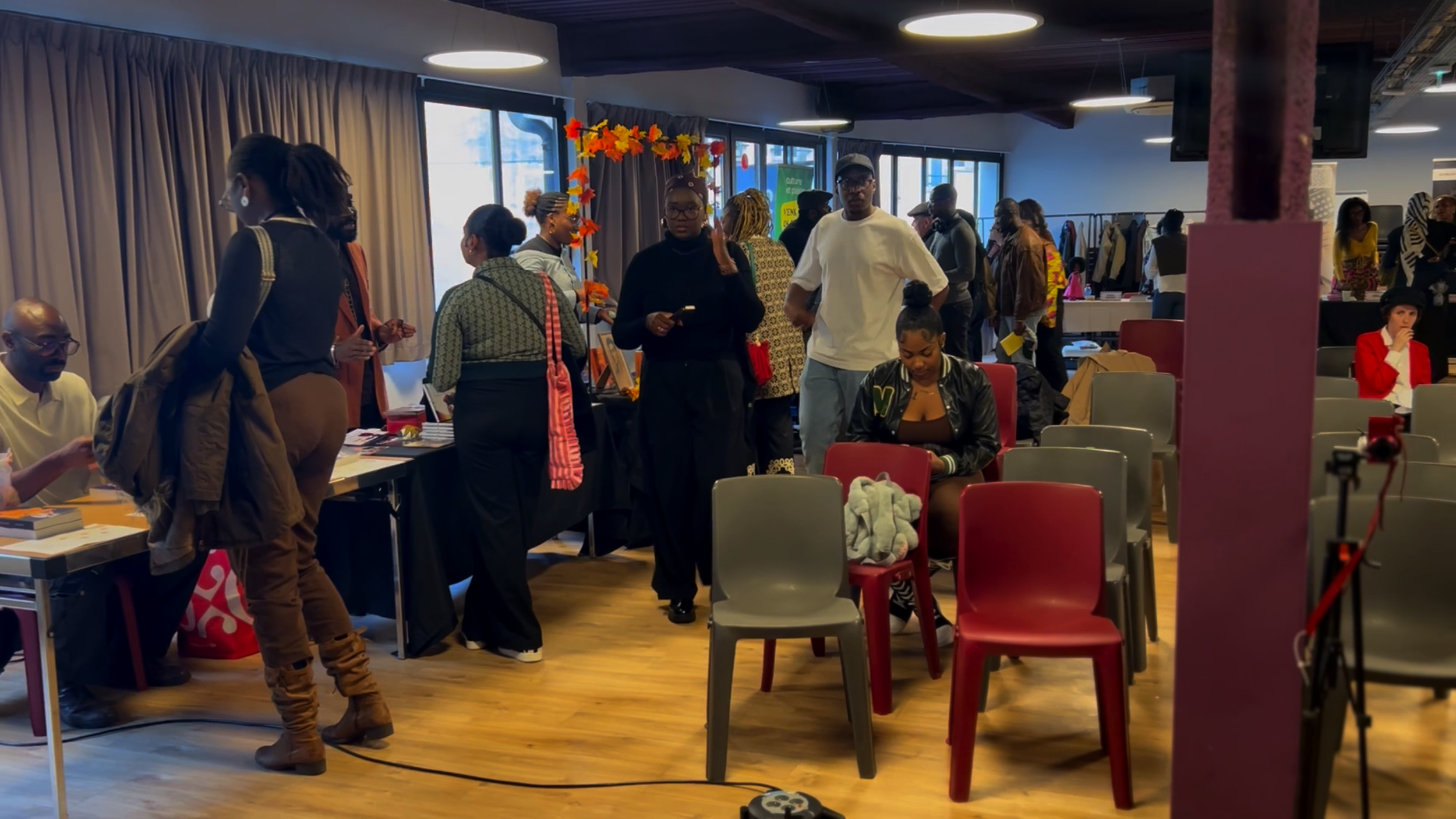 Une salle animée avec des personnes debout et assises, un décor de fête avec guirlandes de feuilles et plusieurs stands ou tables, ambiance chaleureuse et conviviale.