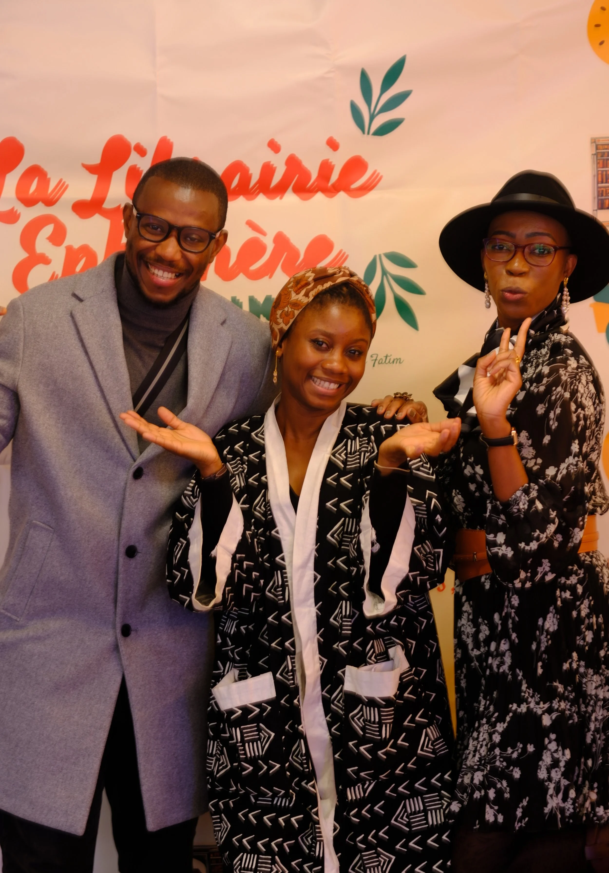 Trois personnes posant avec le sourire devant un fond décoré en orange et vert, avec des livres et une phrase en français.