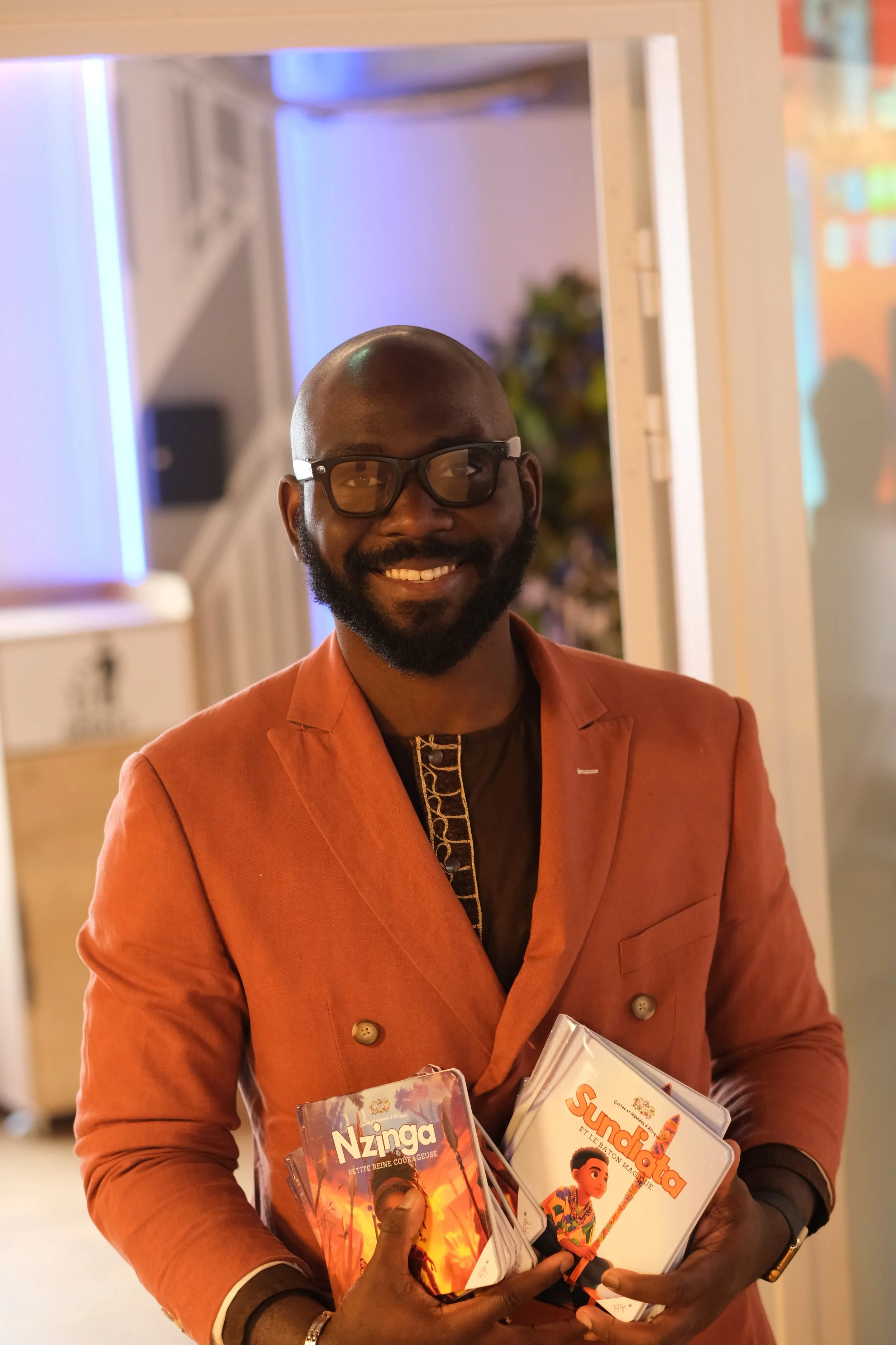 Un homme souriant portant un costume orange, portant des lunettes, tenant des livres pour enfants dans un environnement intérieur