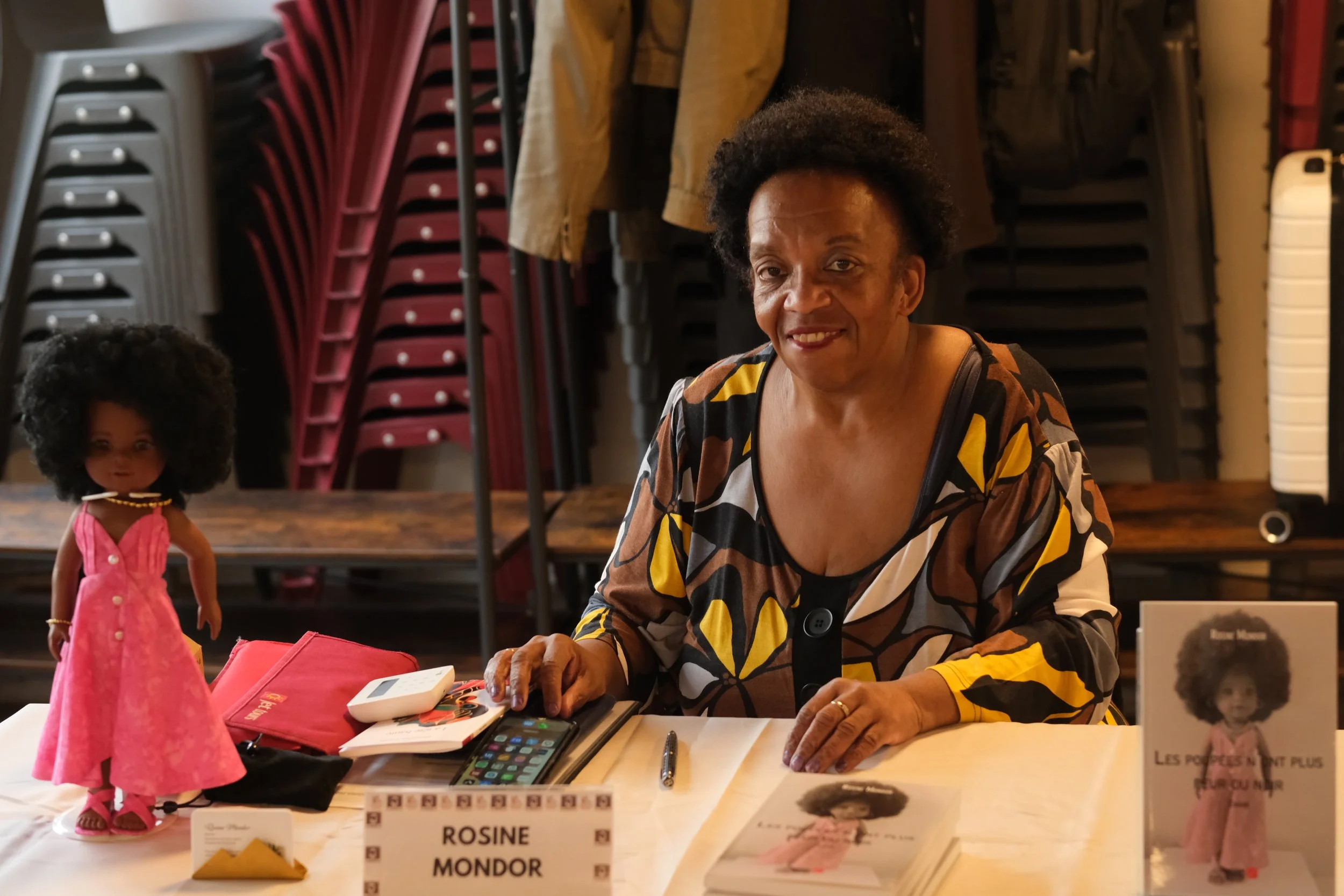 Une femme souriante assise à une table lors d'un événement, avec des chaises empilées en arrière-plan, une poupée ressemblant à une poupée afro, divers objets sur la table, et un panneau avec le nom 'Rosine Mondor'.