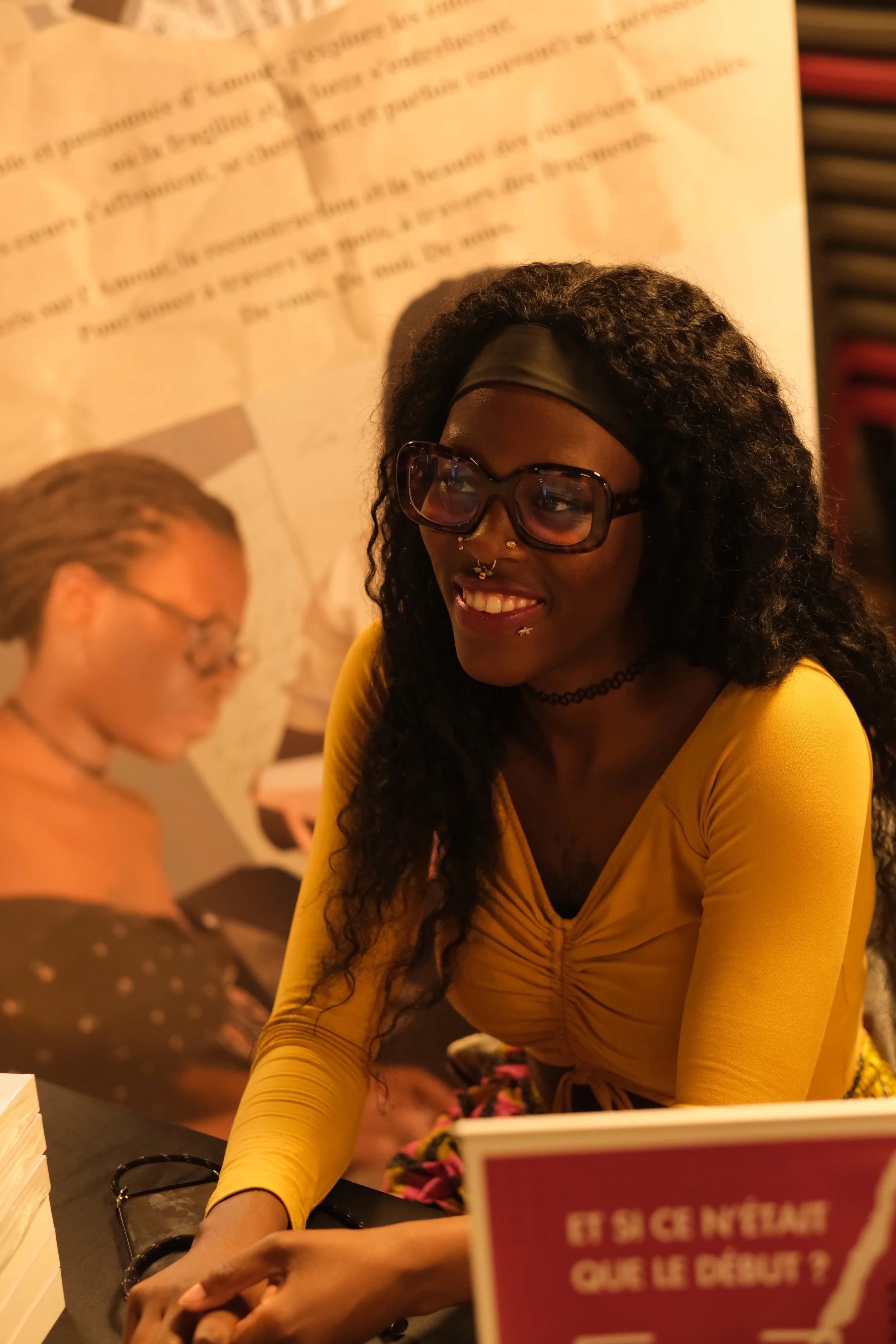 Une femme souriante portant des lunettes et un bandeau noir, vêtue d'un haut jaune, est assise à une table lors d'un événement. En arrière-plan, il y a une affiche avec du texte en français. Une autre personne est également présente, mais floue.