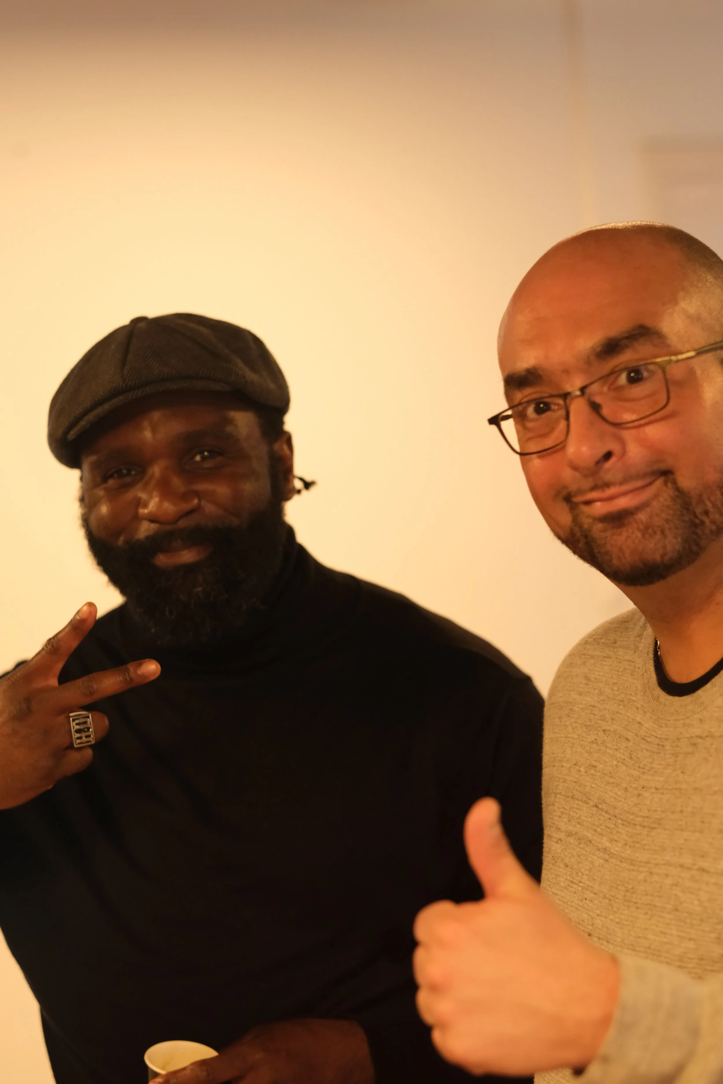Deux hommes prennent un selfie, l'un est noir avec une casquette et la barbe, l'autre est blanc avec des lunettes et un sourire, dans un décor neutre.