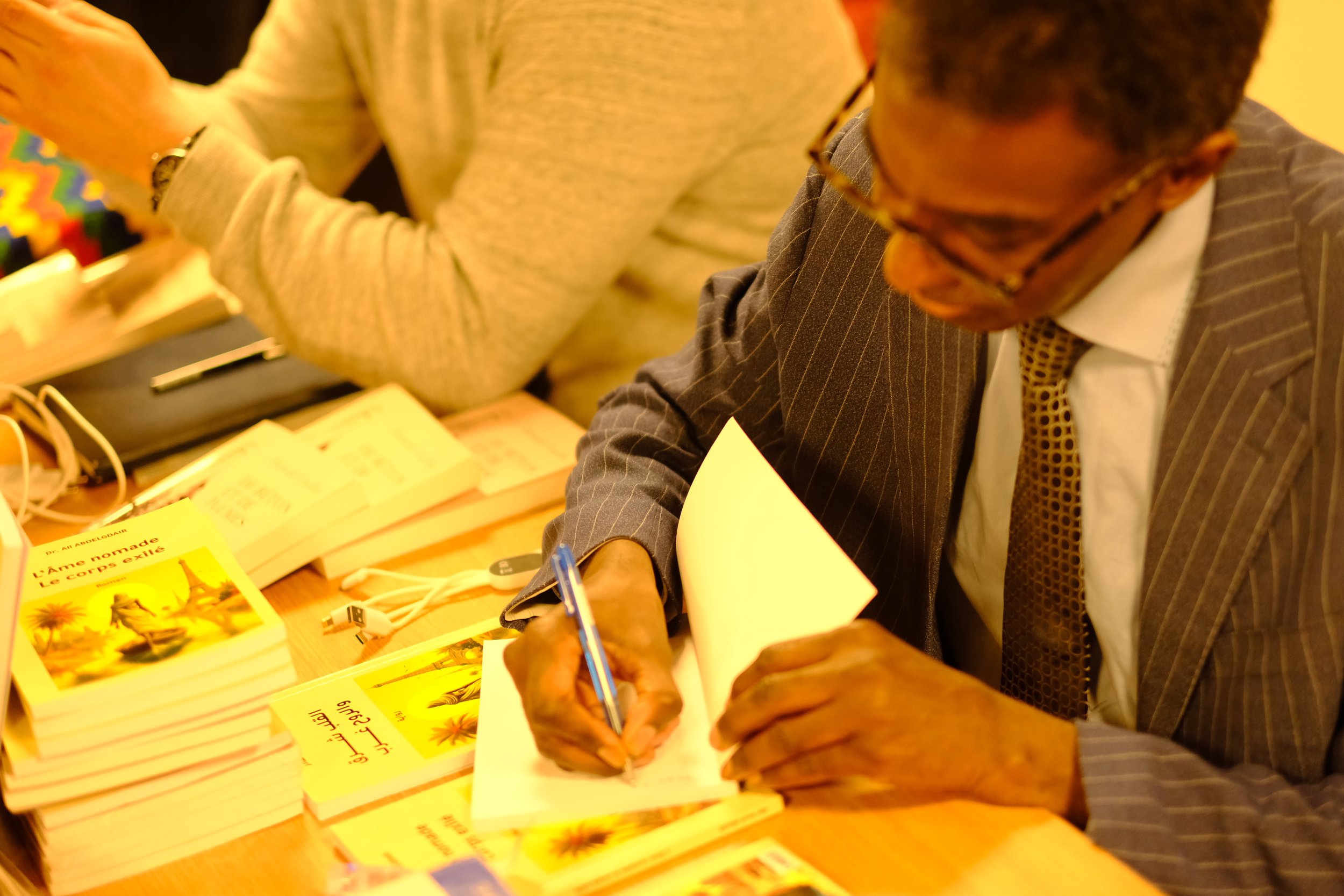 Deux hommes, dont l'un porte un costume à rayures, signent un livre ou une fiche devant une table avec plusieurs livres jaunes empilés. Ils semblent participer à un événement de signature ou de présentation de livres.