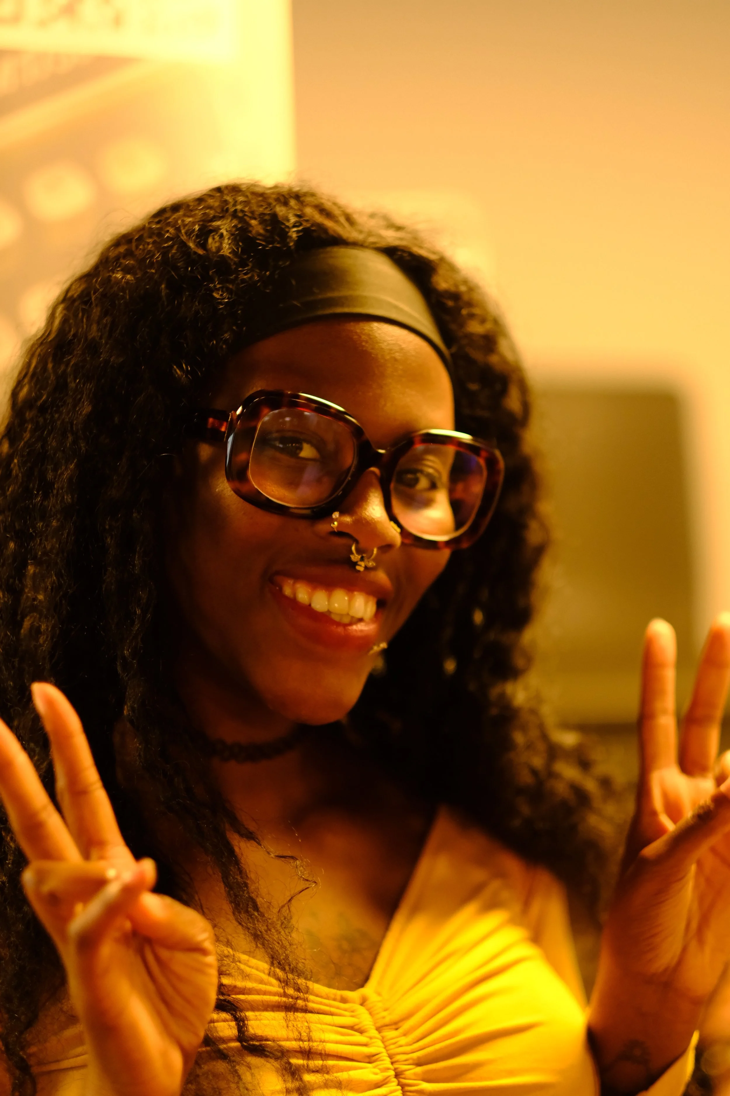 Une femme souriante avec des lunettes grandes, un piercing au nez, en tenue jaune, faisant un signe de paix avec les doigts.
