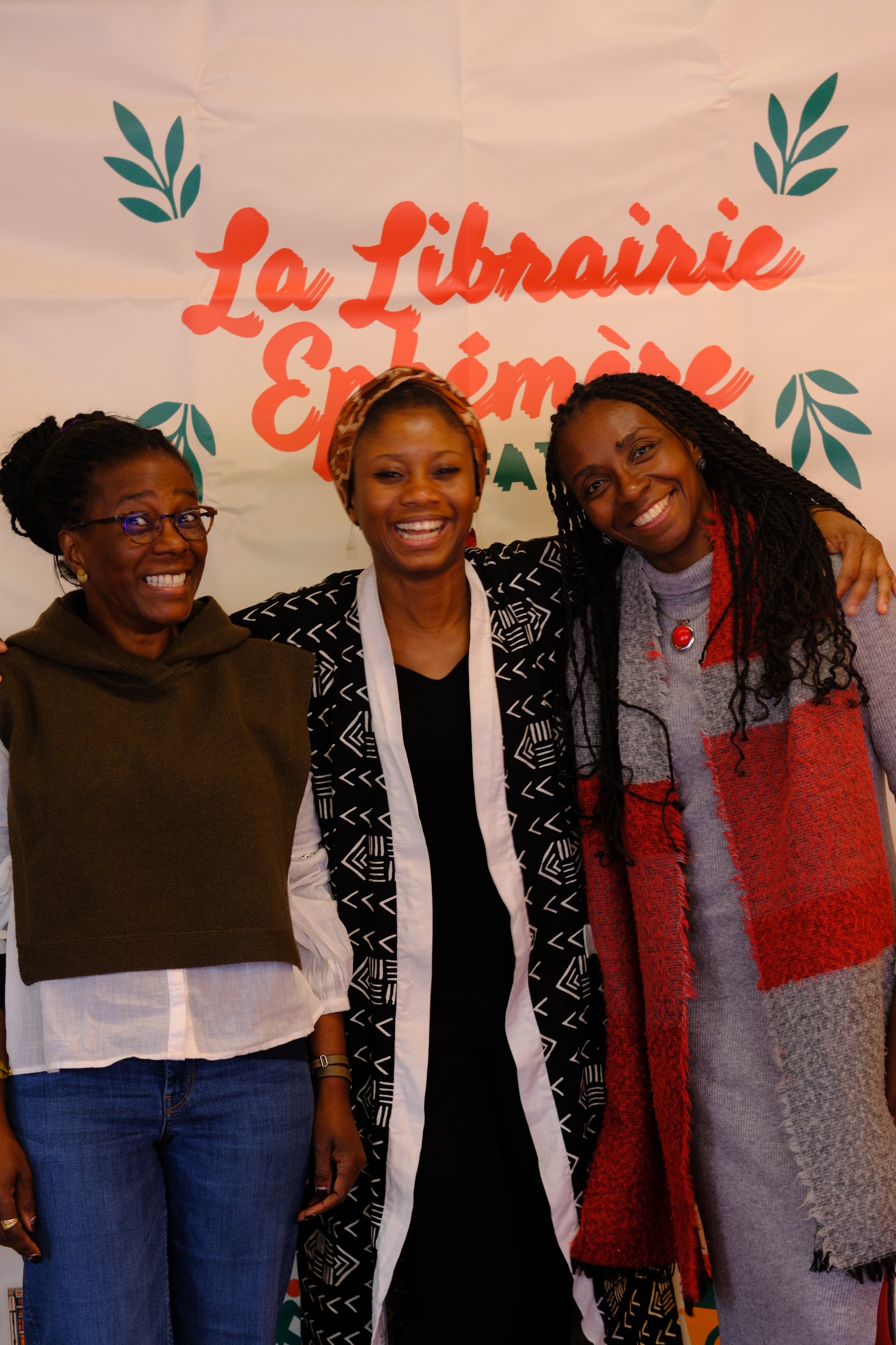 Trois femmes souriantes posant devant un fond avec le texte 'La Libération En Faisant' et des décorations vertes, en intérieur.