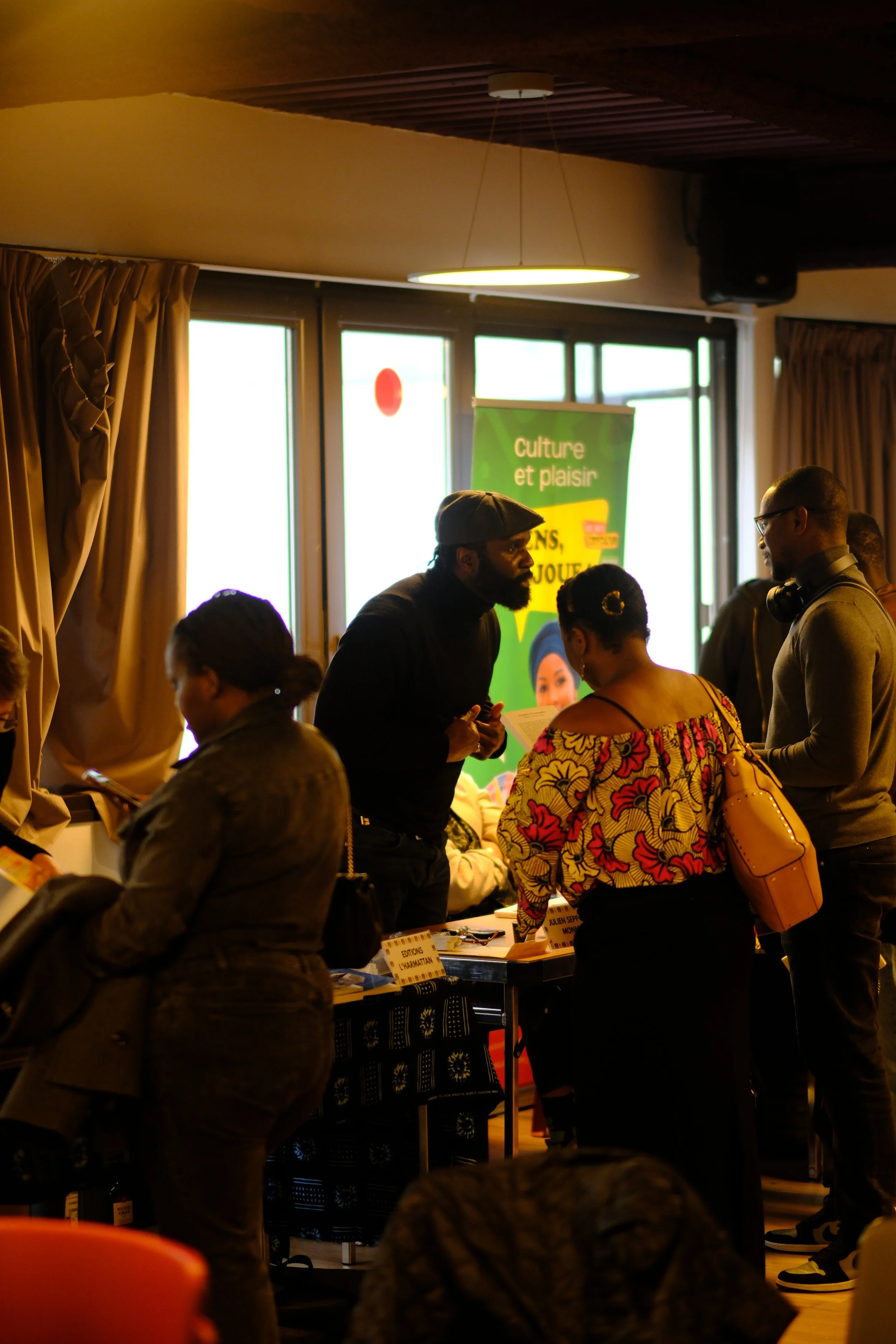 Groupe de personnes à une table lors d'un événement à l'intérieur, avec un fond affichant un panneau vert. Les personnes discutent, l'une d'elles tient un document.