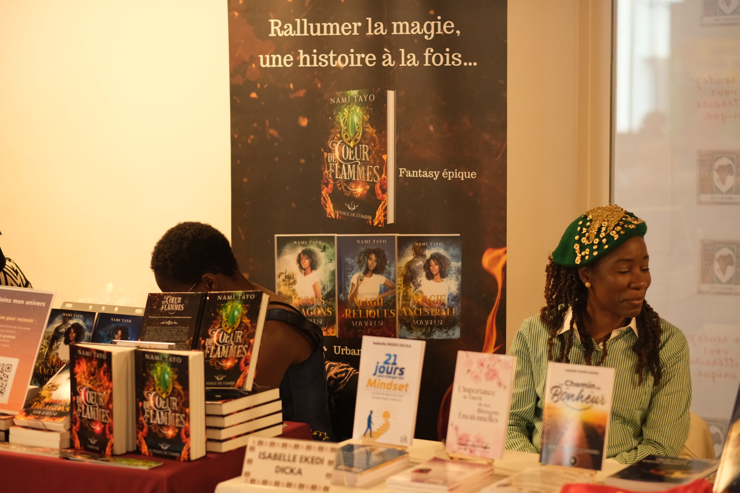 Photographie d'un stand de livres avec une grande affiche promotionnelle pour un roman intitulé 'Coeur de Flammes' de Nami Tayo. Plusieurs exemplaires du livre sont exposés sur la table, et on voit deux femmes derrière le stand, dont l'une porte un c