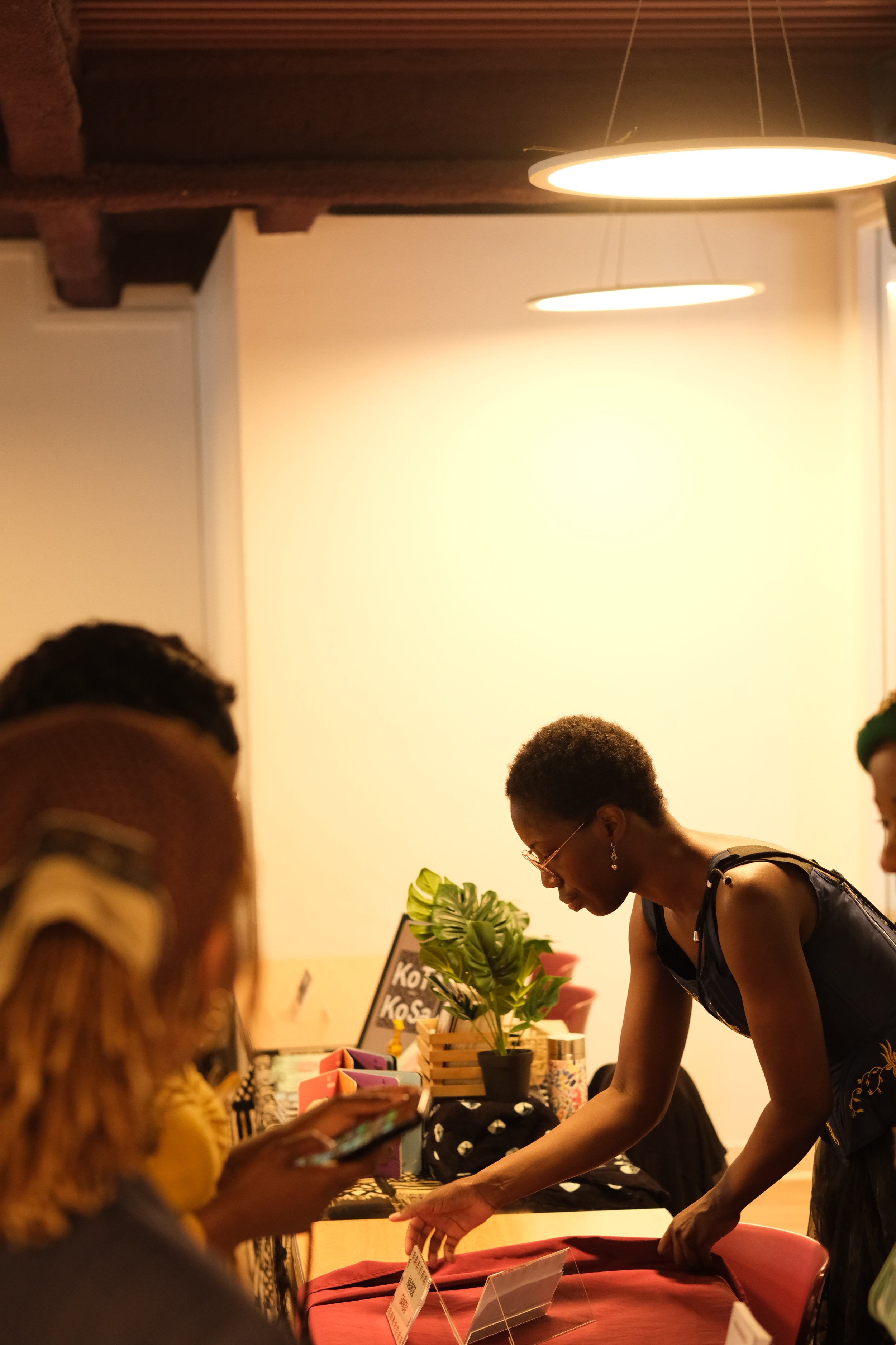 Une femme interagit avec des visiteurs lors d'un évènement, exposant probablement des produits ou des œuvres d'art. L'ambiance est chaleureuse avec un éclairage ambiant et des plantes décoratives sur la table.