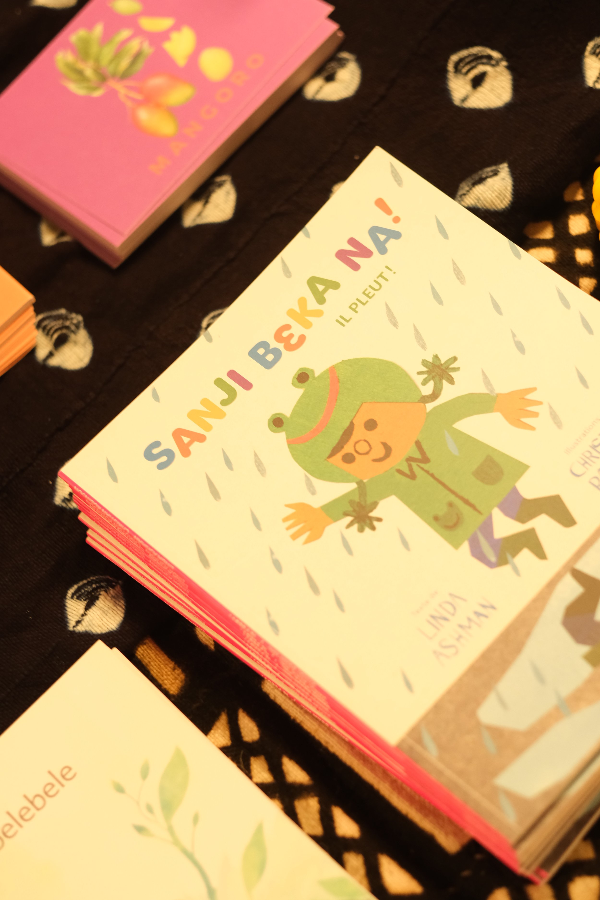 La photo montre une pile de livres pour enfants avec une couverture colorée illustrant une grenouille et un titre en français. La pile est posée sur un tissu noir avec un motif de feuilles blanches.