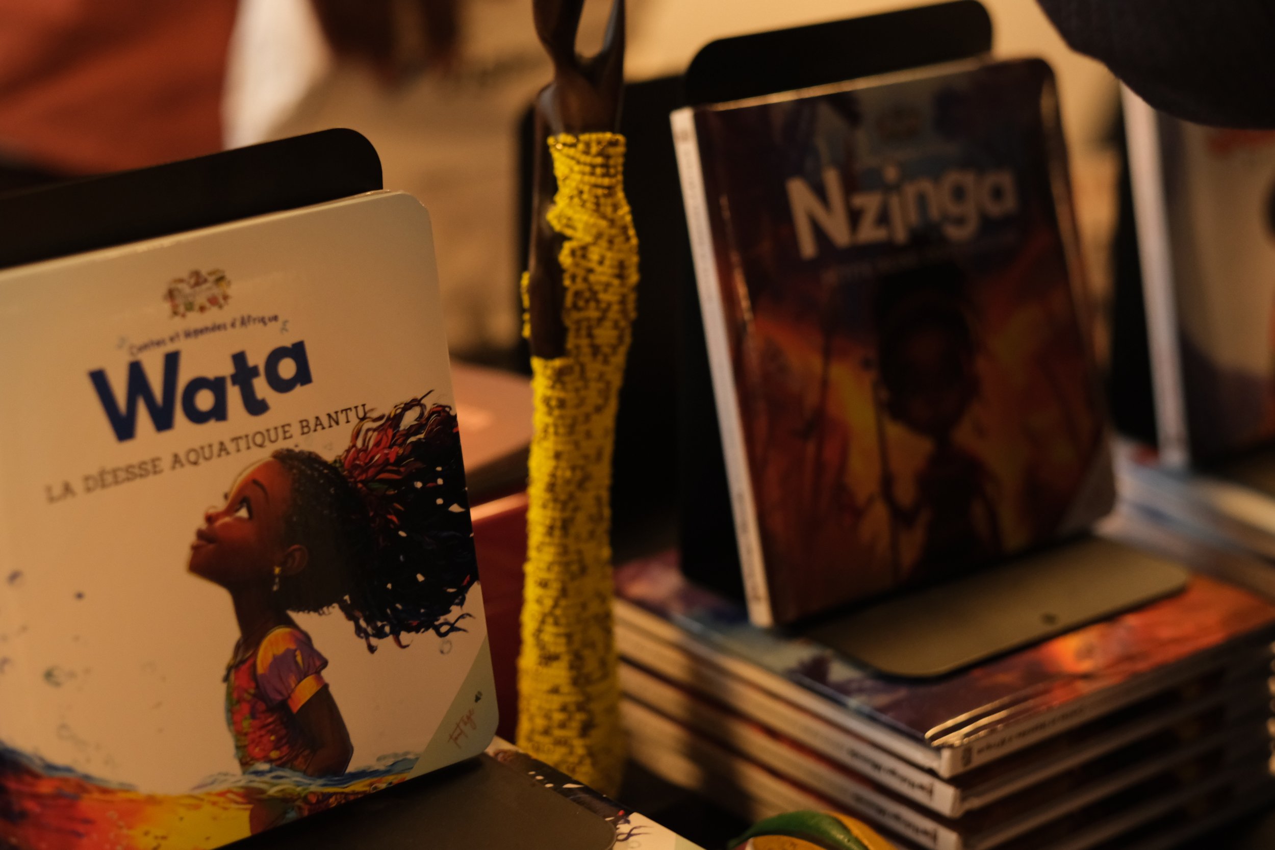 Livre avec couverture représentant une femme noire avec des cheveux crépus, coloration chaude et illustration d'une scène africaine, placé à gauche. À droite, une pile de bandes dessinées ou cartes avec un marque-page jaune attaché. En arrière-plan, 