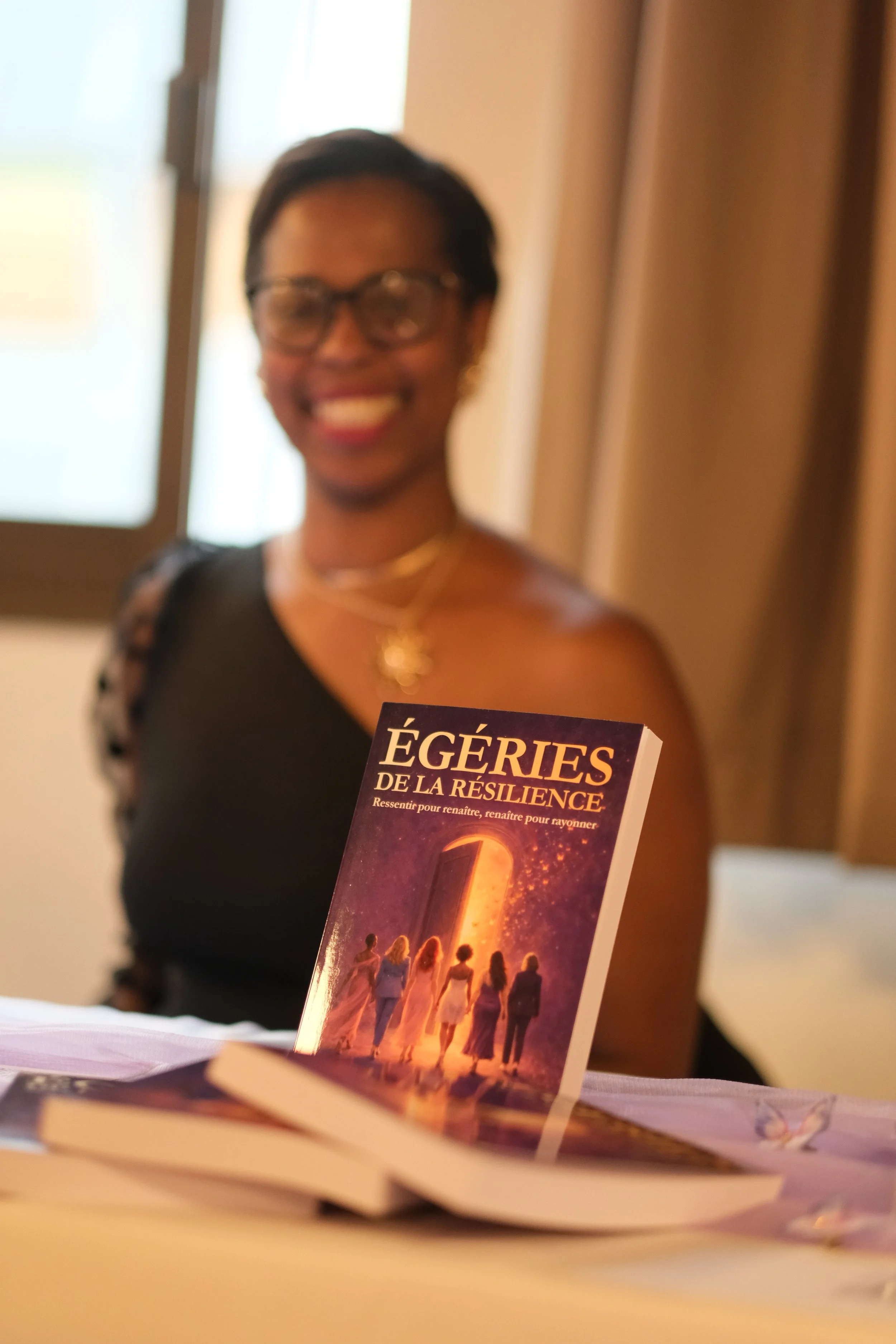 Une femme souriante avec des lunettes tient un livre intitulé "ÉGÉRIES DE LA RÉSILIENCE" devant une fenêtre. Le fond est flou et la photo est légèrement floue.