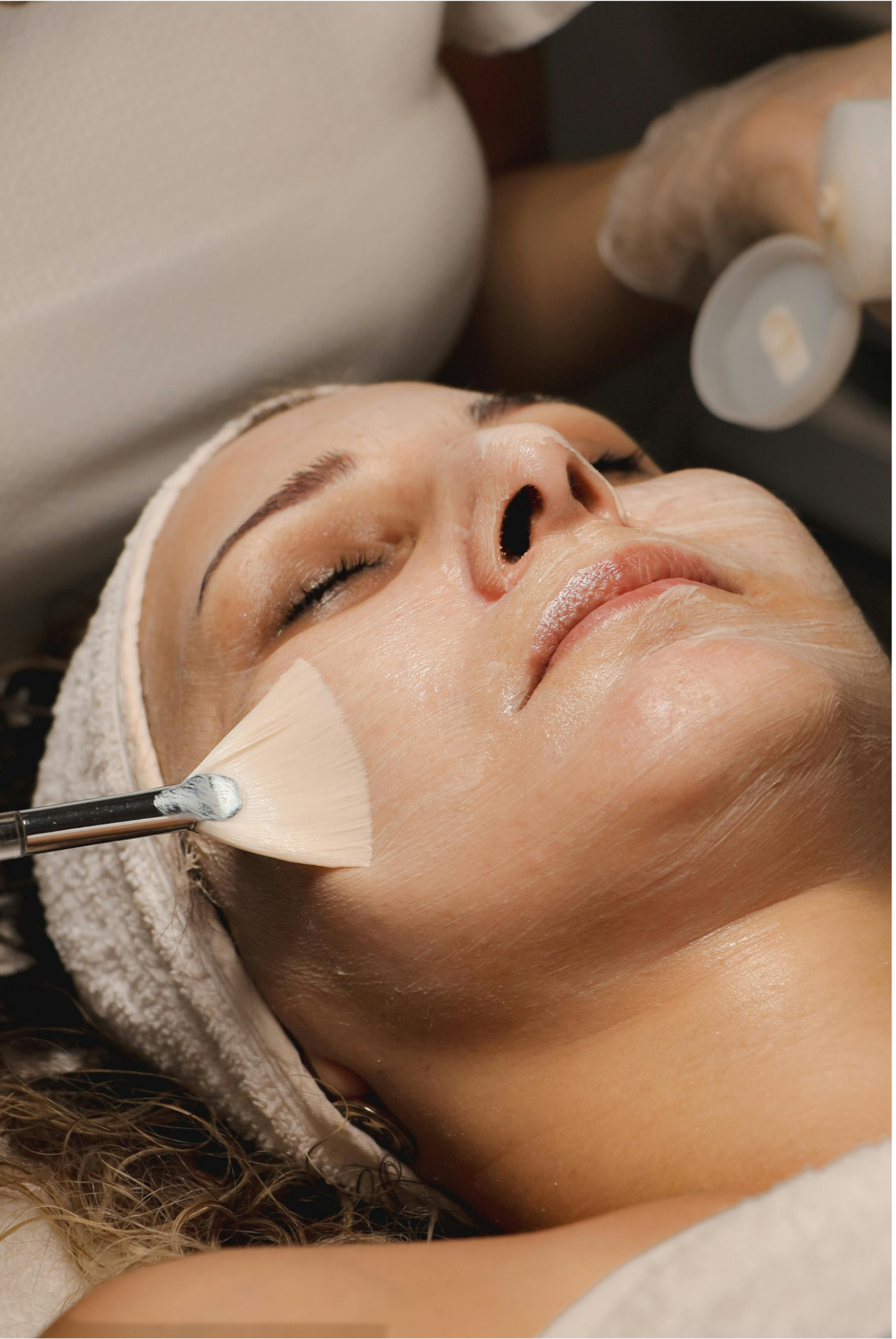 A woman lying down with eyes closed, receiving a facial treatment with a brush applying a product on her cheek.